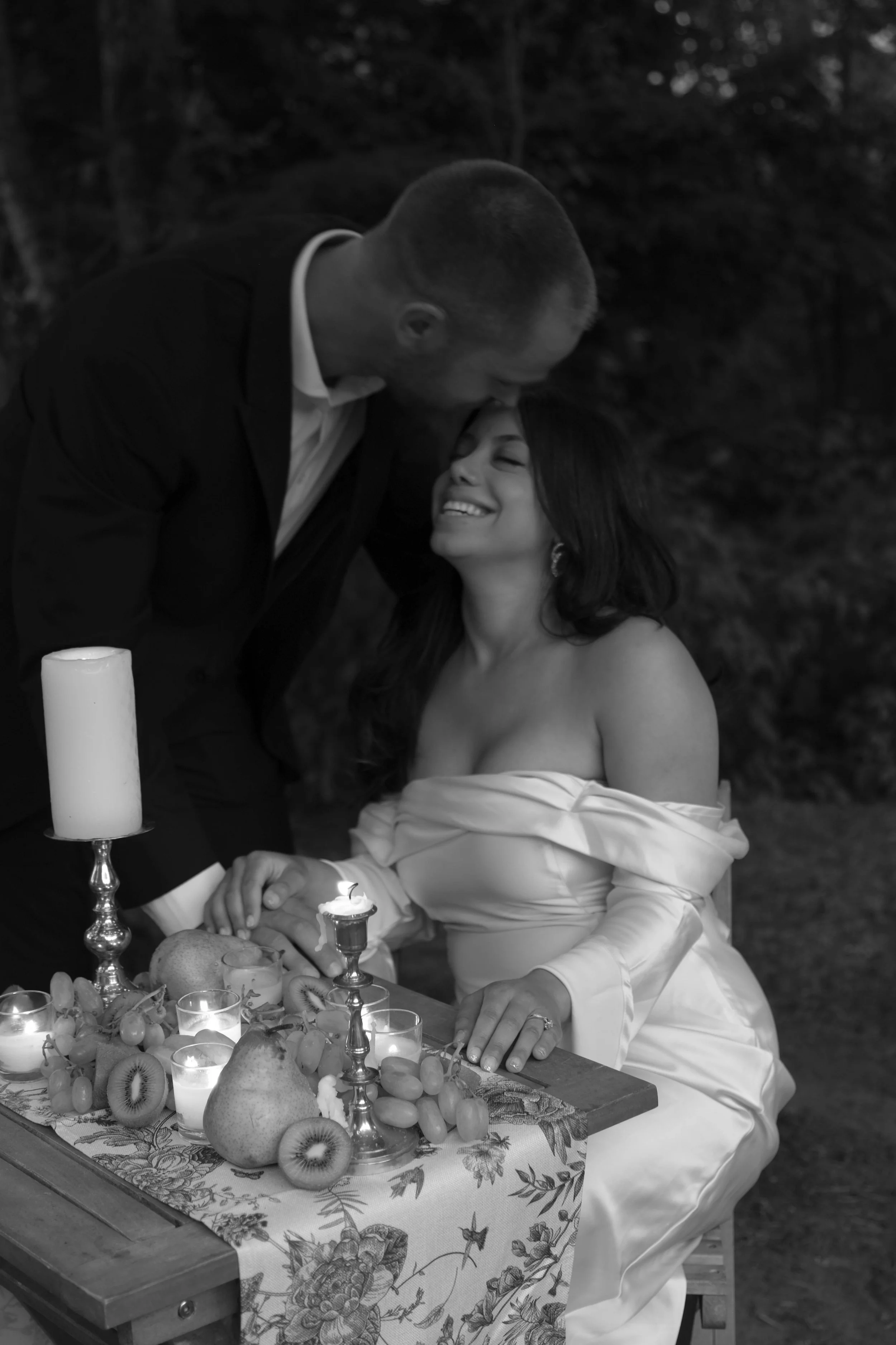 A black and white photo of a joyful woman in a strapless dress sitting at a table with an assortment of fruits and candles. A man in a suit leans over her, kissing her forehead, creating an intimate moment amid an outdoor setting at dusk or nighttime.