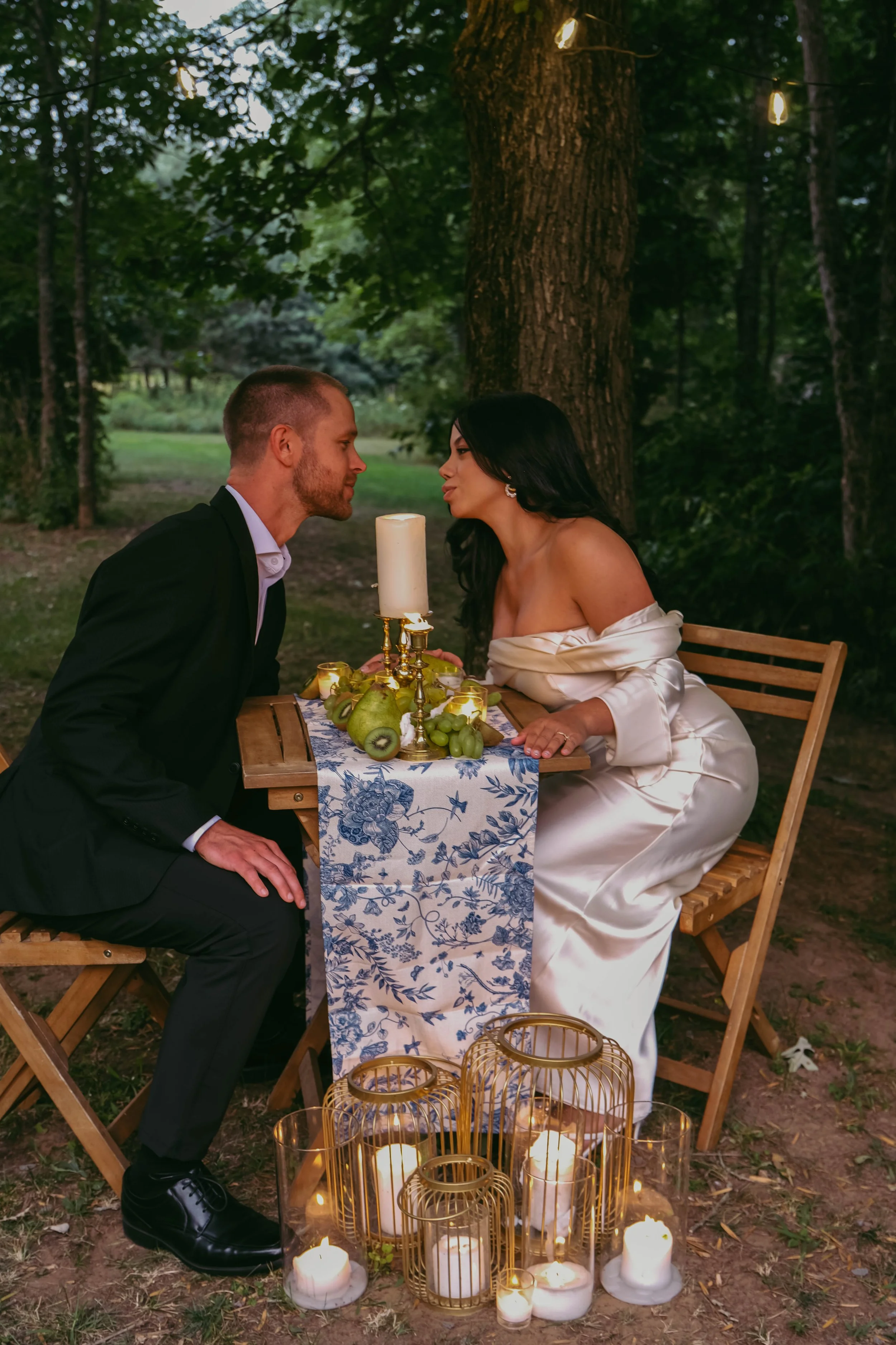 Potsdam, NY Photographer A couple dressed in formal attire sitting at a table outdoors under a tree at dusk, with candles and string lights creating a romantic setting.