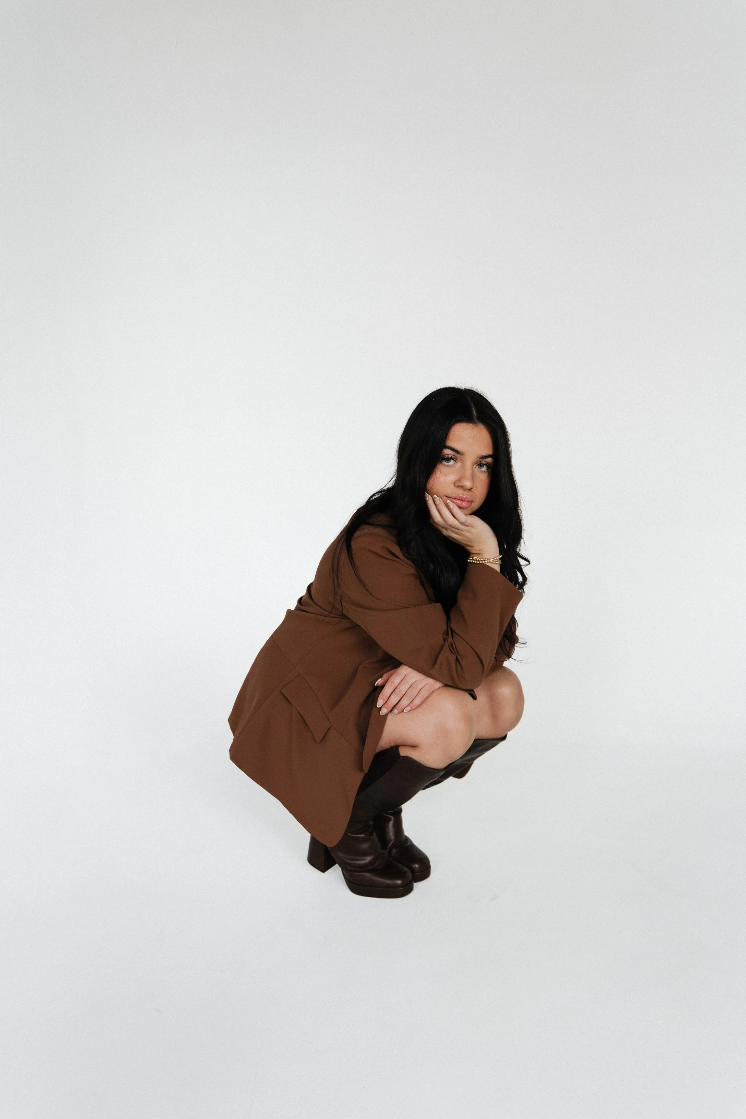 Syracuse, NY Branding photos, A woman with long black hair crouching down against a plain white background, wearing a brown jacket, dark boots, and minimal jewelry.