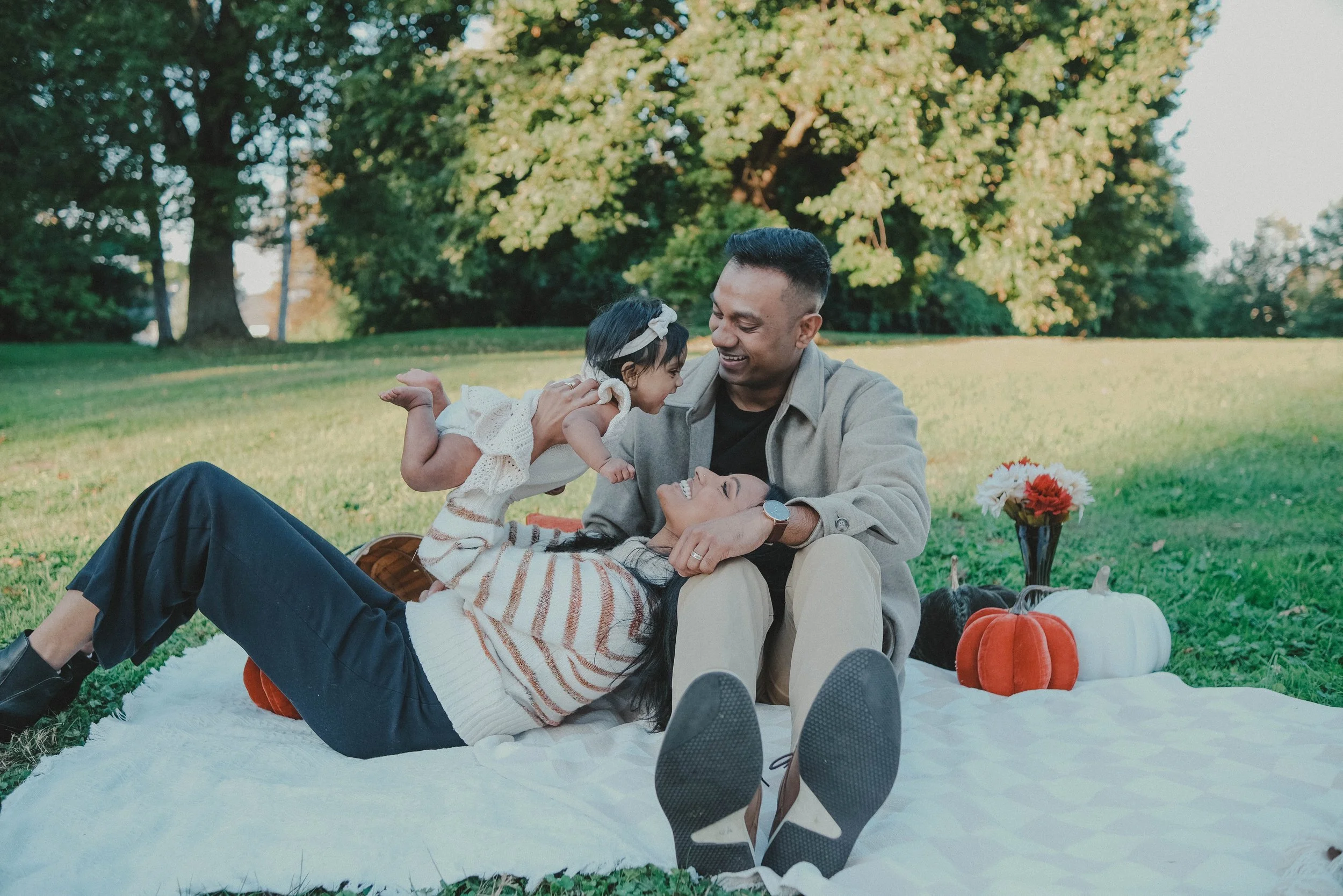Burnet Park, Syracuse NY A family enjoying a picnic outdoors on a sunny day, with a woman lying down on the blanket, a man sitting beside her, and a young girl leaning towards them. There are pumpkins and a vase of flowers on the blanket.