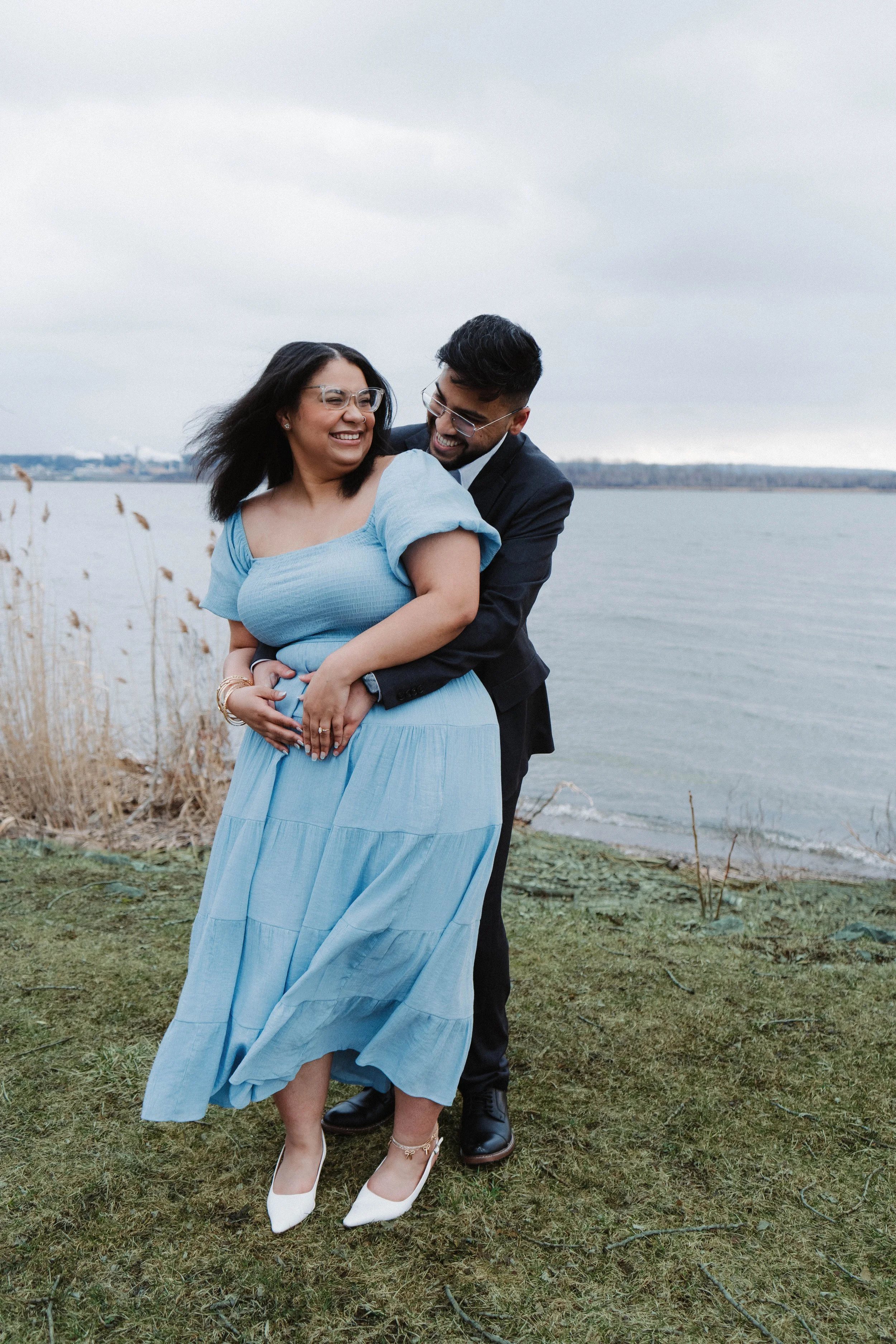 A couple smiling and embracing outdoors near a body of water, with grass and reeds in the background, on a cloudy day.