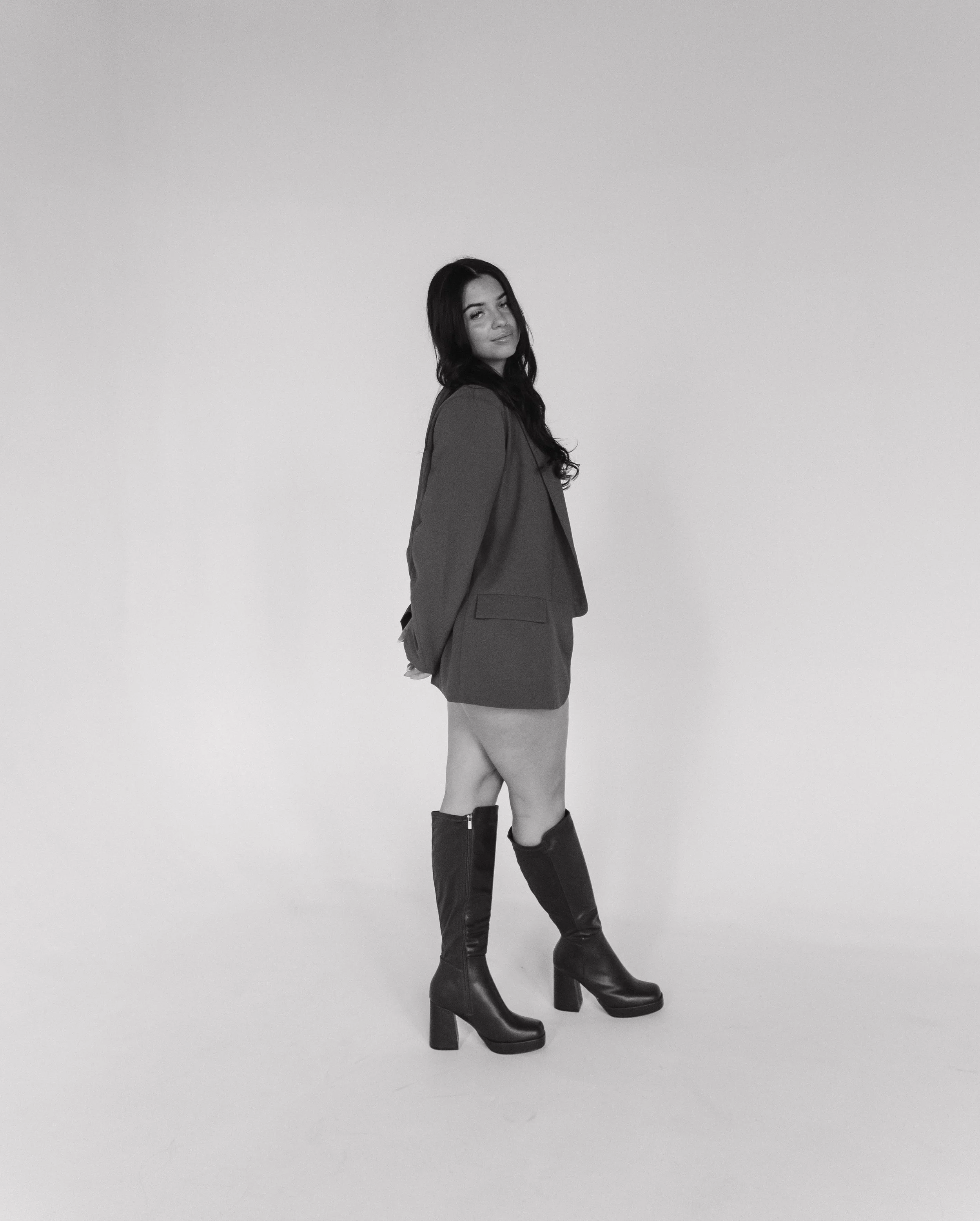 A woman with long dark hair wearing a blazer, shorts, and high-heeled boots, standing against a plain white background.