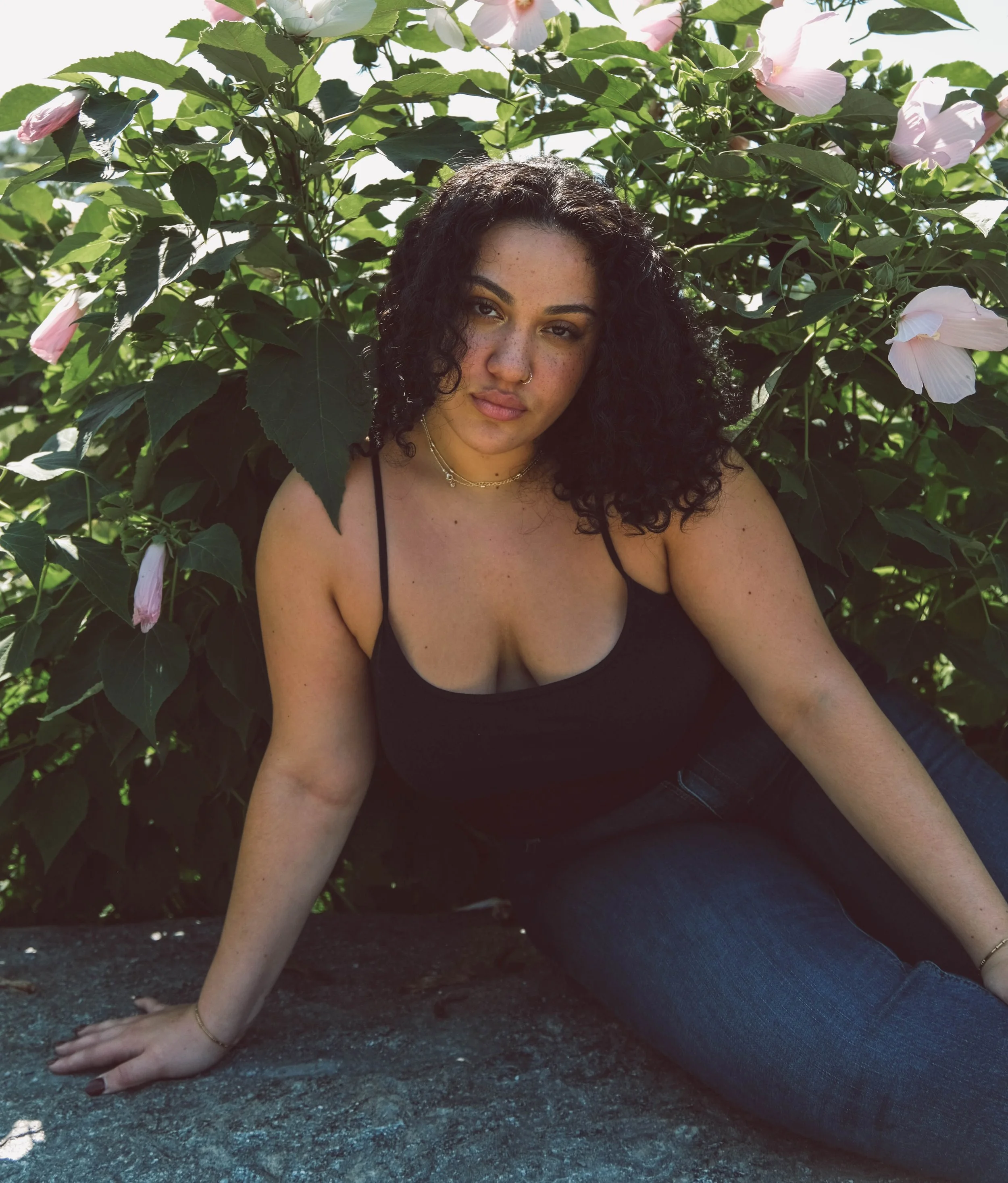 Central Park, A woman with curly dark hair, wearing a black spaghetti strap top and dark jeans, sitting on a rock in front of green leafy plants with pink and white flowers, outdoors in sunlight.