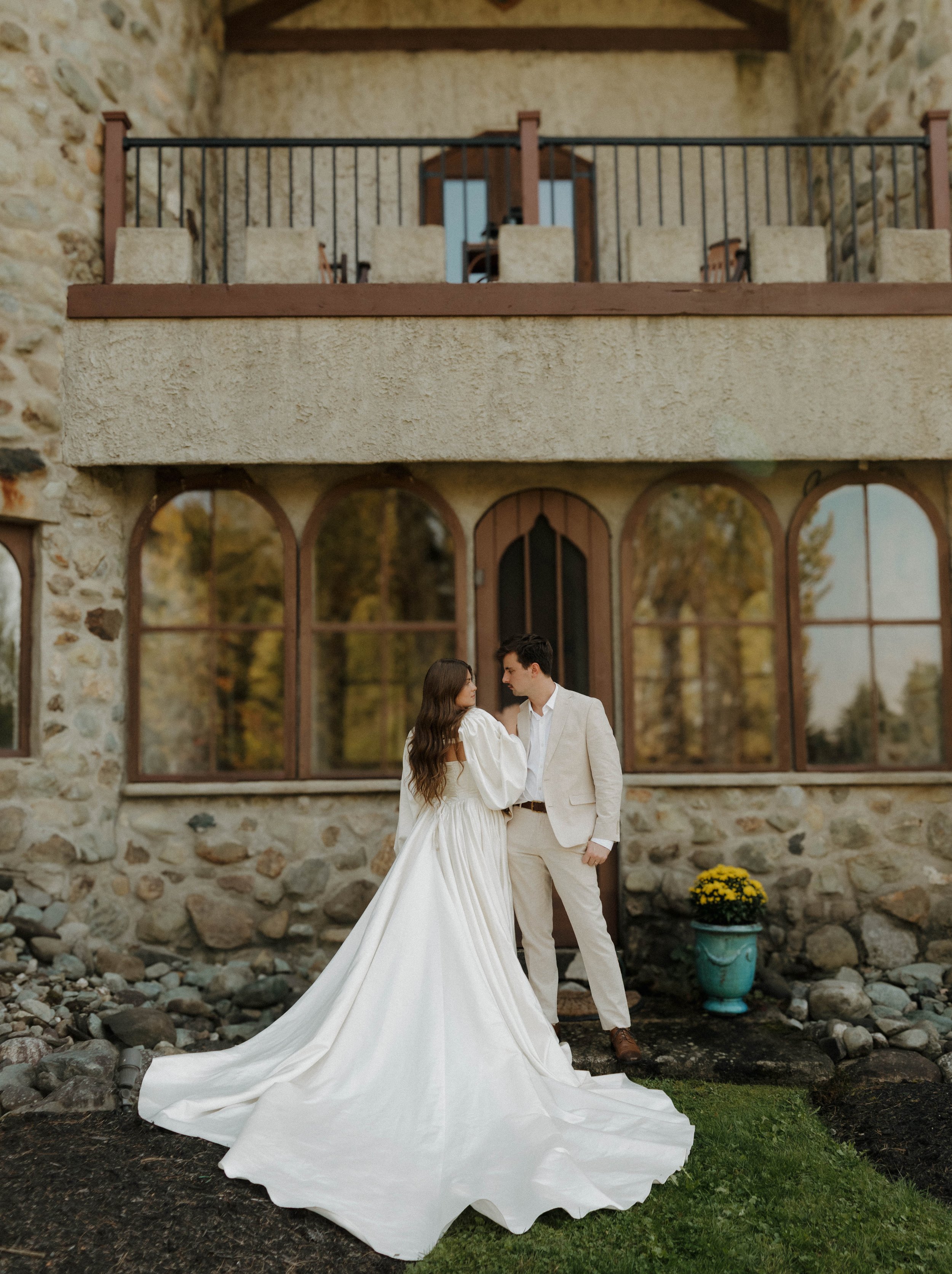 Bride in a white wedding gown and groom in a cream suit stand close together outside a stone building with large arched windows, surrounded by trees and boulders.