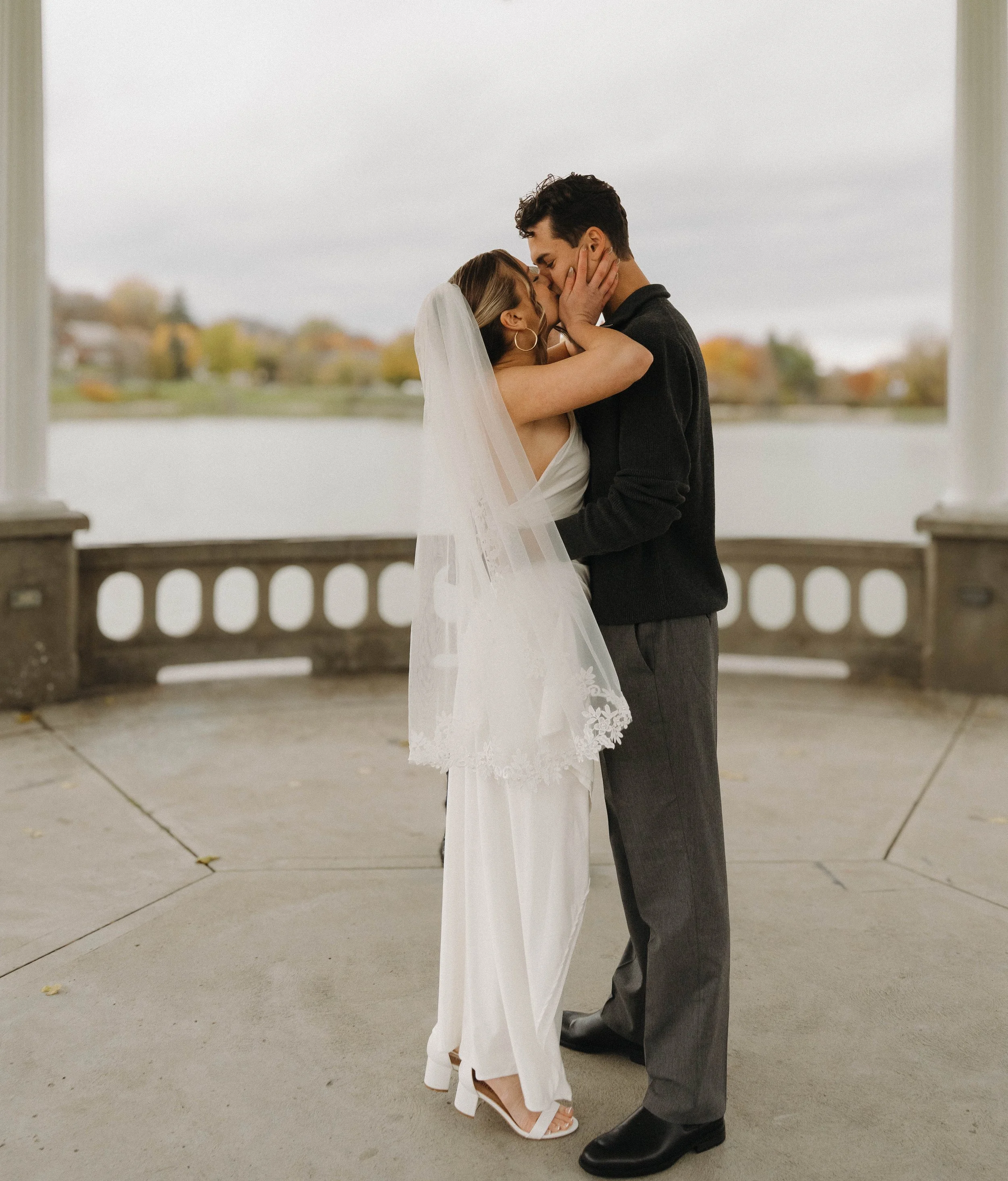 A bride and groom embrace and kiss on a pavilion overlooking a lake with fall foliage in the background.