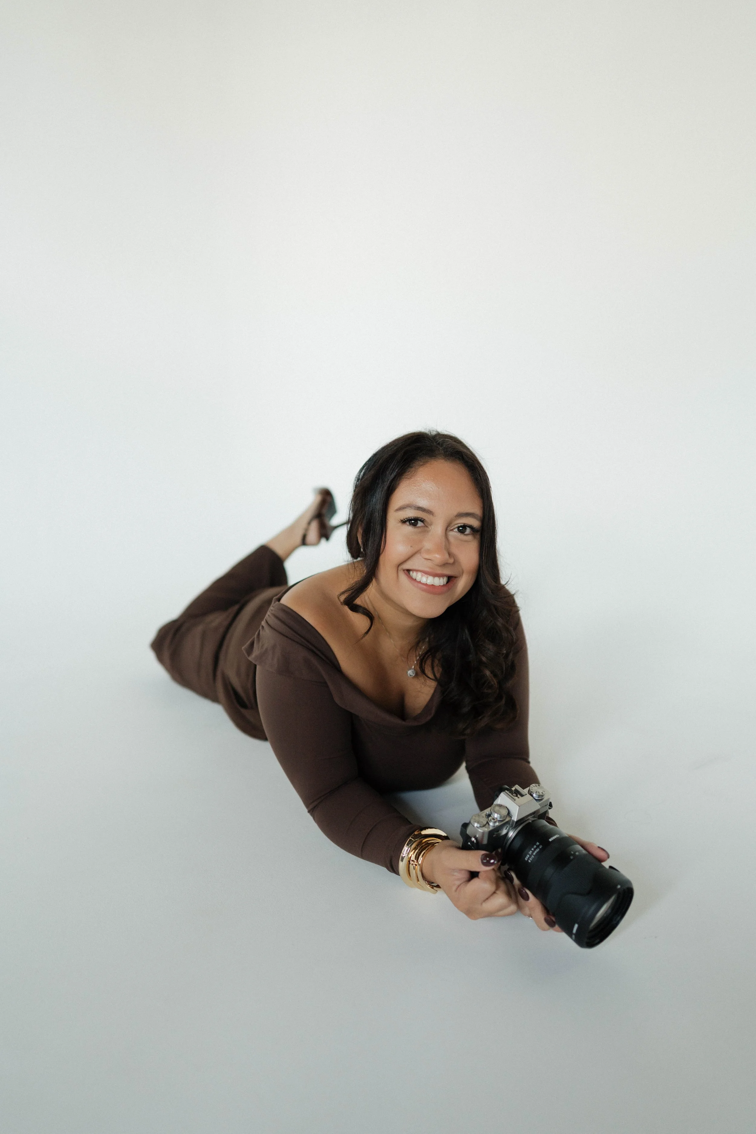 Woman lying on white surface, smiling, holding camera, wearing brown dress and gold jewelry.