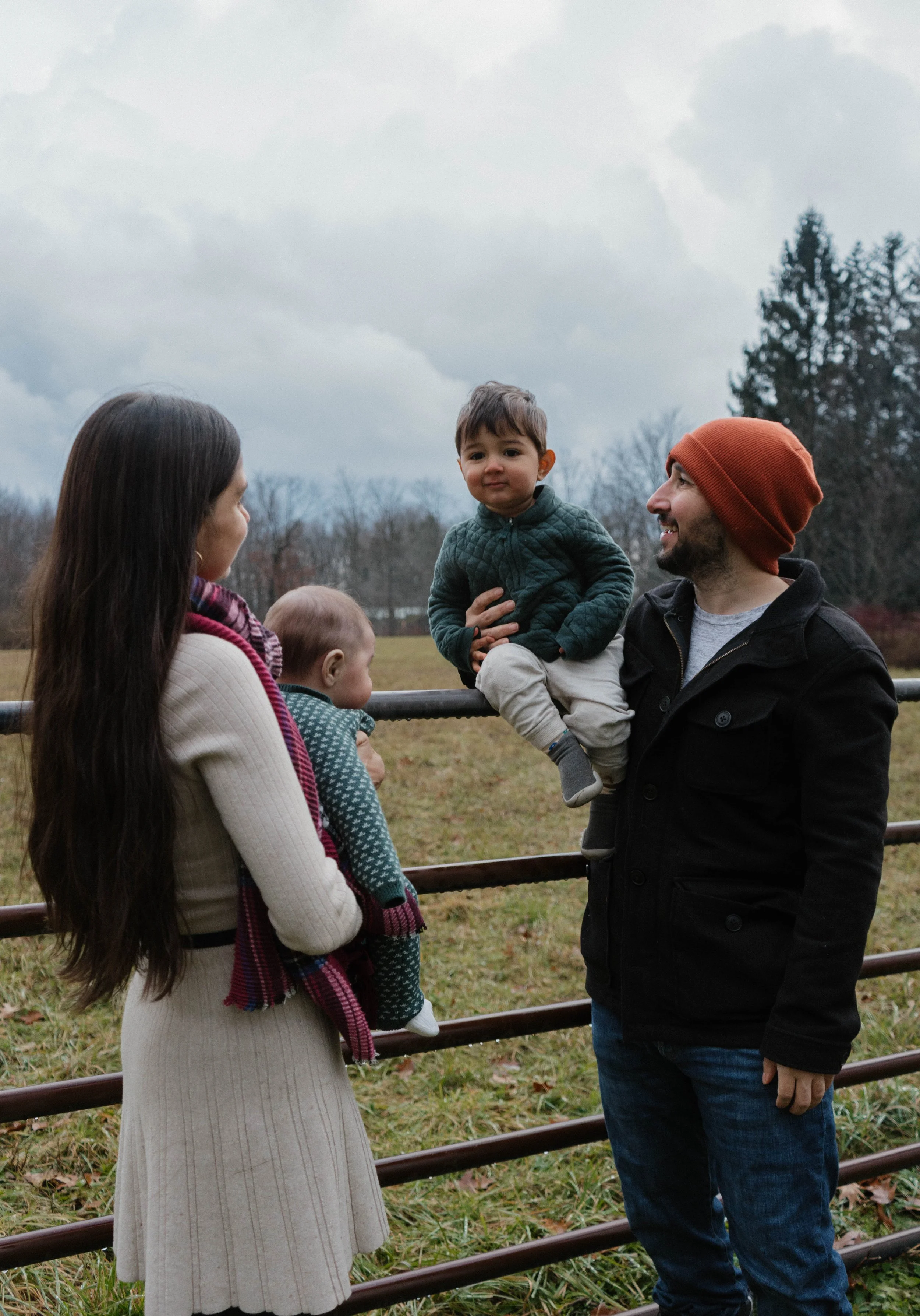 A family of four standing outdoors near a metal fence, with a grassy field and trees in the background. The father, wearing a red beanie and black jacket, is holding a young boy, and the mother, with long dark hair, is holding a baby while talking to the father.