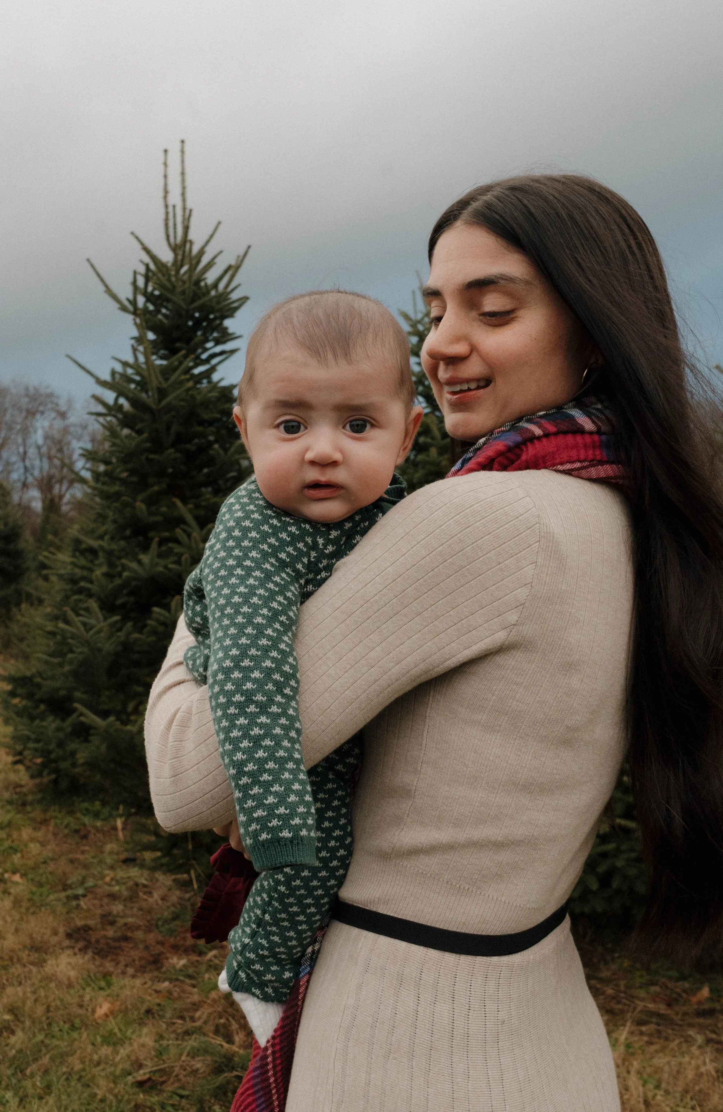 Page's Christmas Tree Farm, Syracuse, NY A woman holding a young child outdoors with Christmas trees in the background, wearing a beige dress with a black belt and a red plaid scarf, and the child wearing a green patterned sweater.