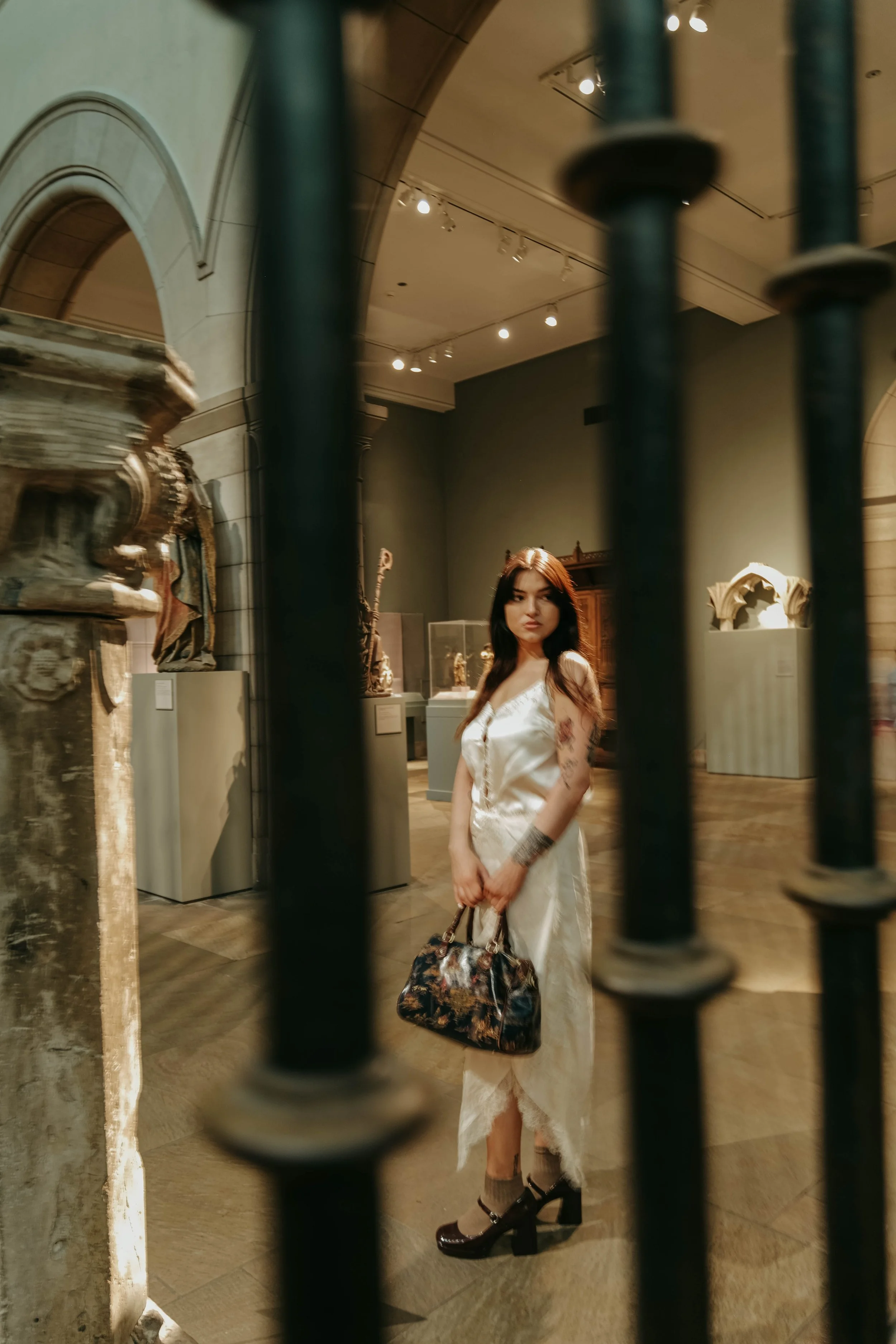 The Met Museum, A woman with long dark hair, wearing a white dress, standing behind a black metal railing in an art or history museum, holding a patterned handbag, surrounded by sculptures and historical artifacts.