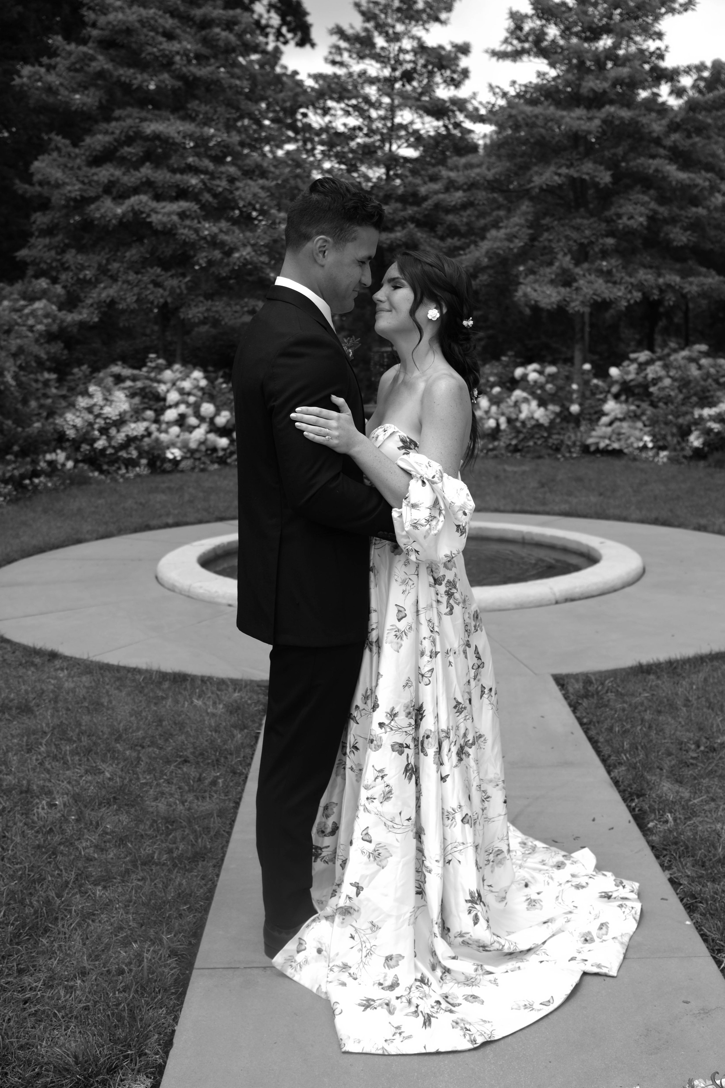 A black and white photo of a couple dressed in wedding attire embracing in a garden. The groom is wearing a dark suit and tie, and the bride is in a strapless floral wedding gown. They are standing on a pathway near a landscaped fountain with trees and flowers in the background.