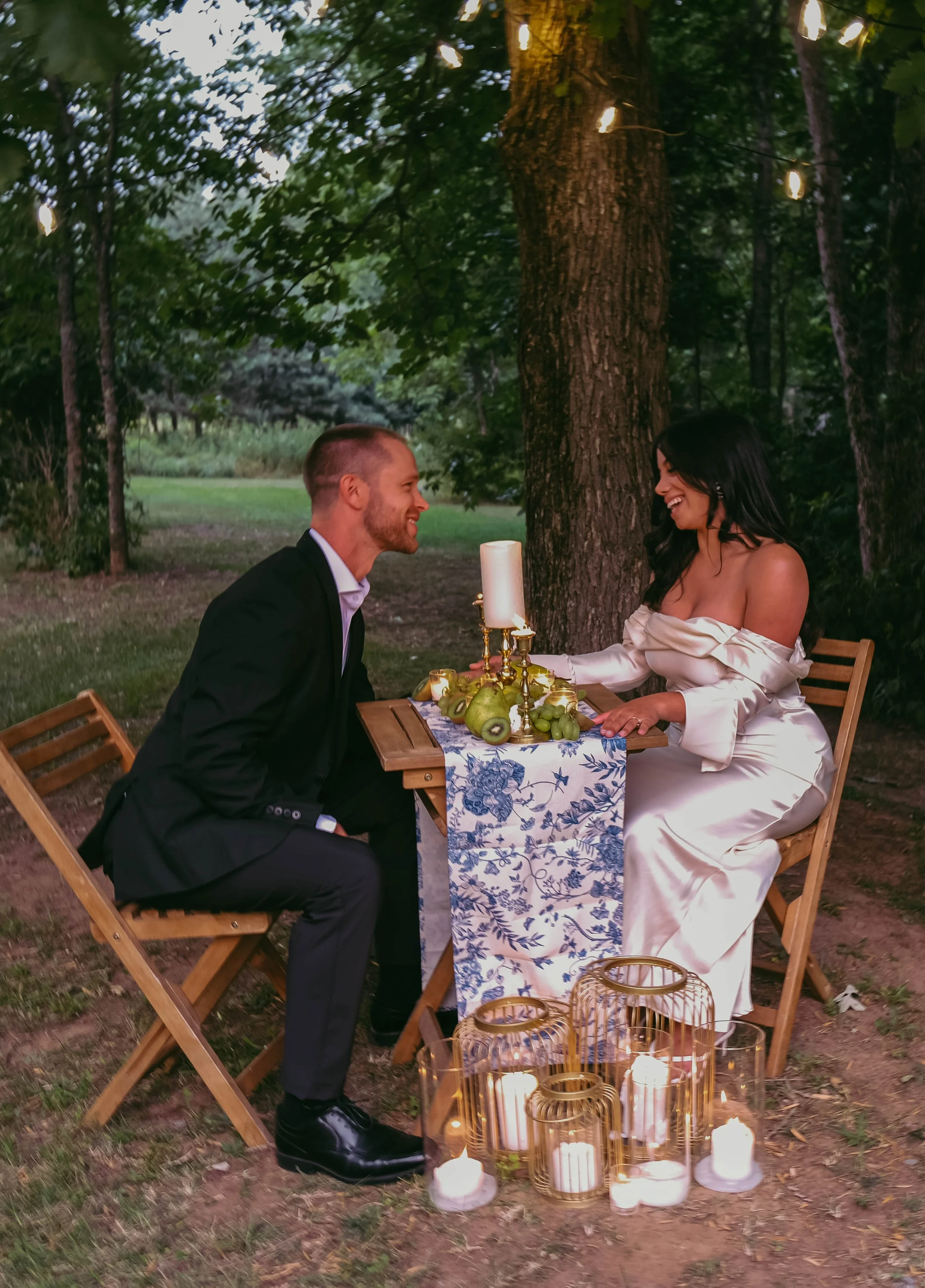 Potsdam, NY Photographer A couple sitting at a romantic outdoor dinner table with candles and greenery, sharing a joyful moment.