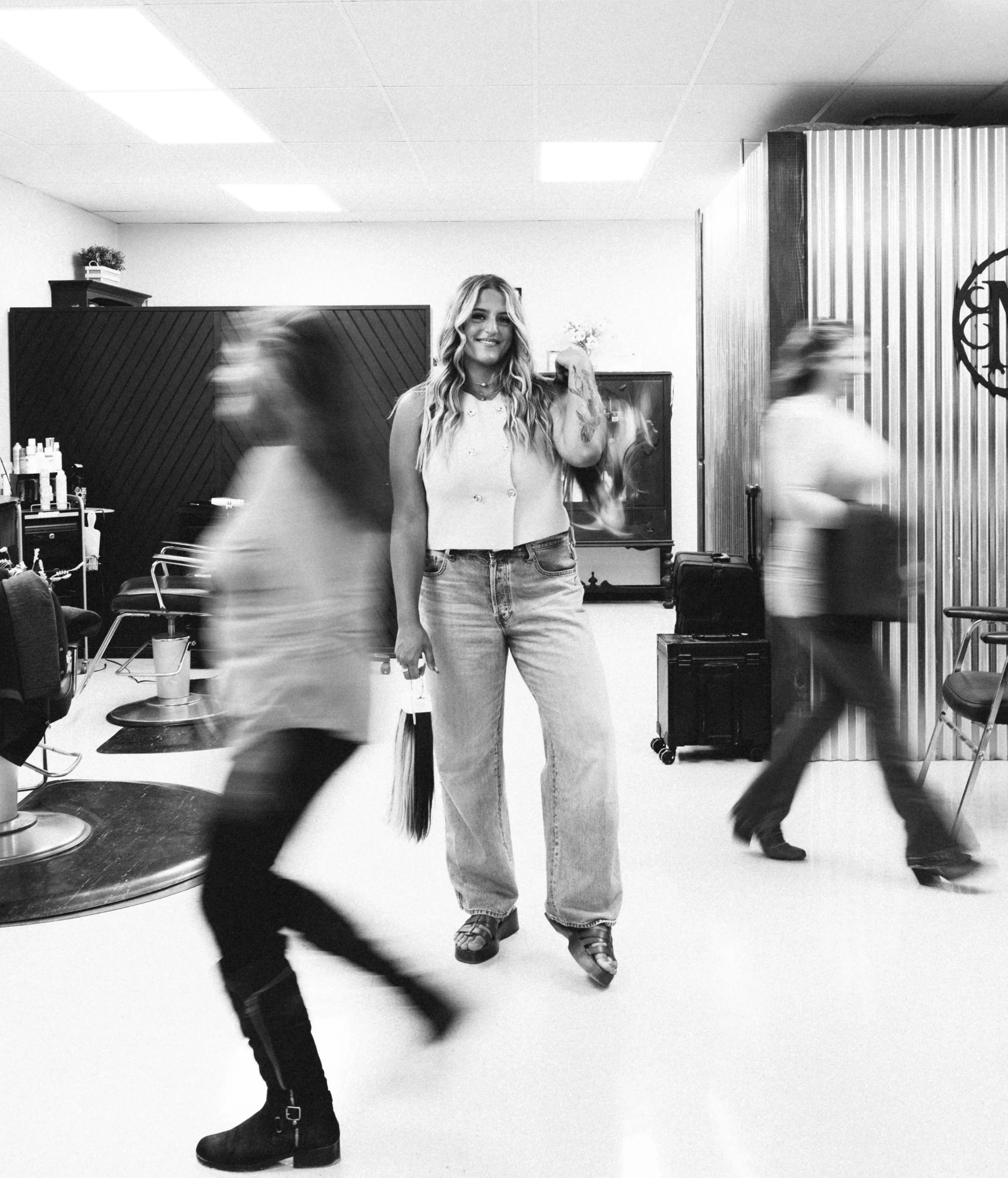 A young woman with long wavy hair smiling in a salon among two people walking past her, one in a sleeveless top and jeans, the other in a T-shirt and pants, with hair and faces blurred from motion.