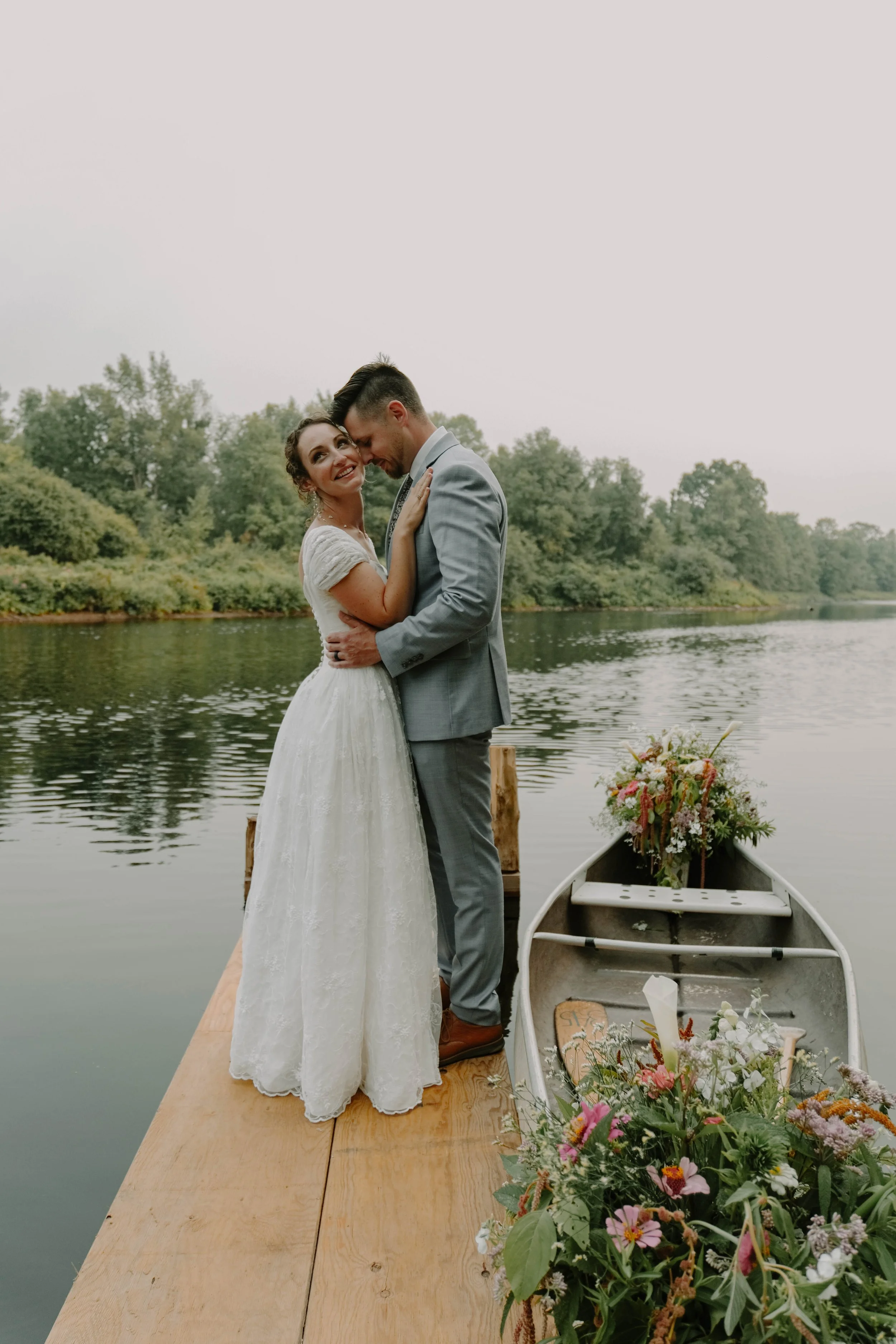 A newlywed couple in wedding attire embracing on a wooden dock by the water, with a canoe decorated with flowers nearby and lush greenery in the background.
