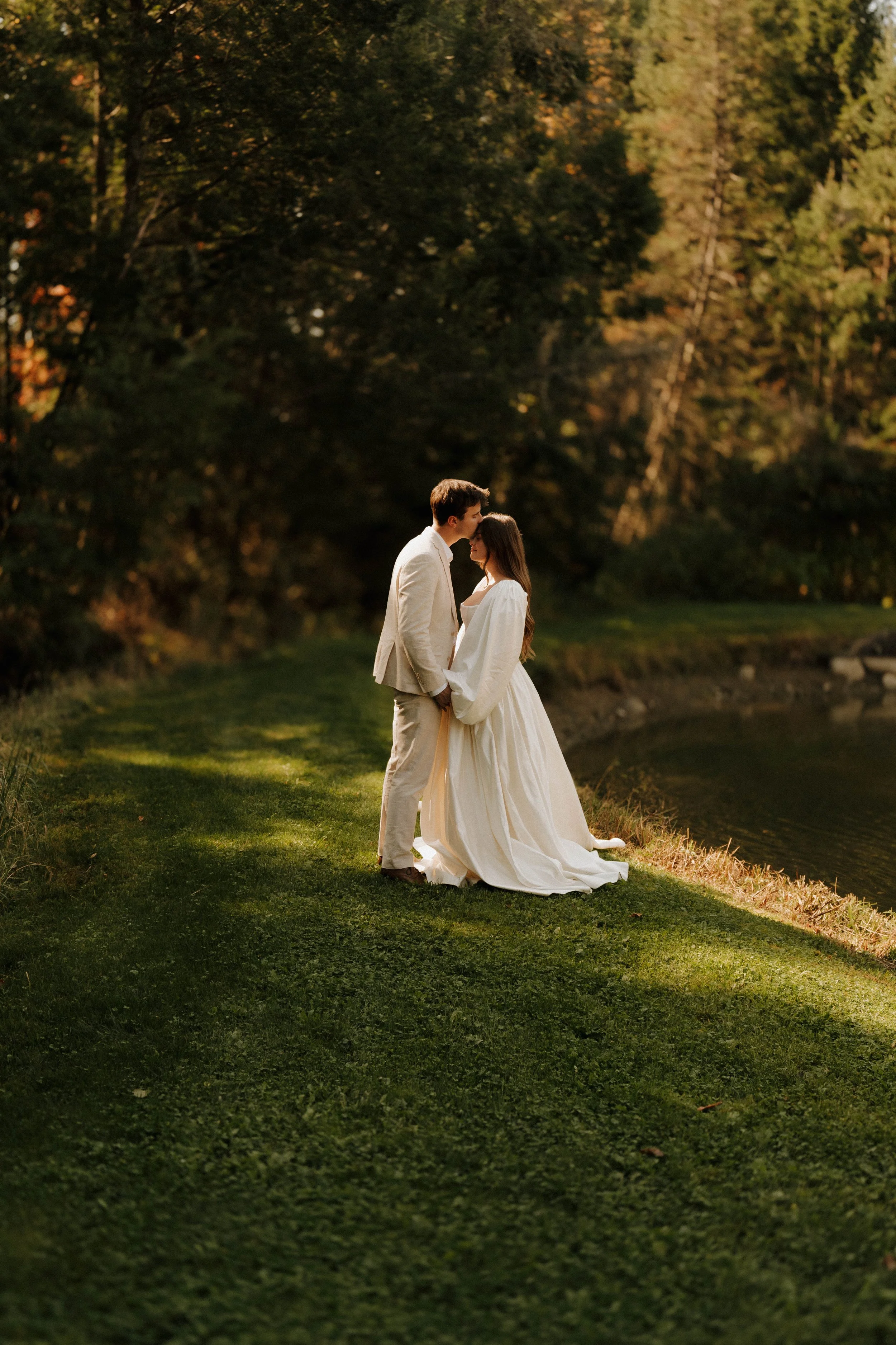 A couple in wedding attire standing by a river at sunset, holding hands and touching foreheads, surrounded by trees with autumn foliage.