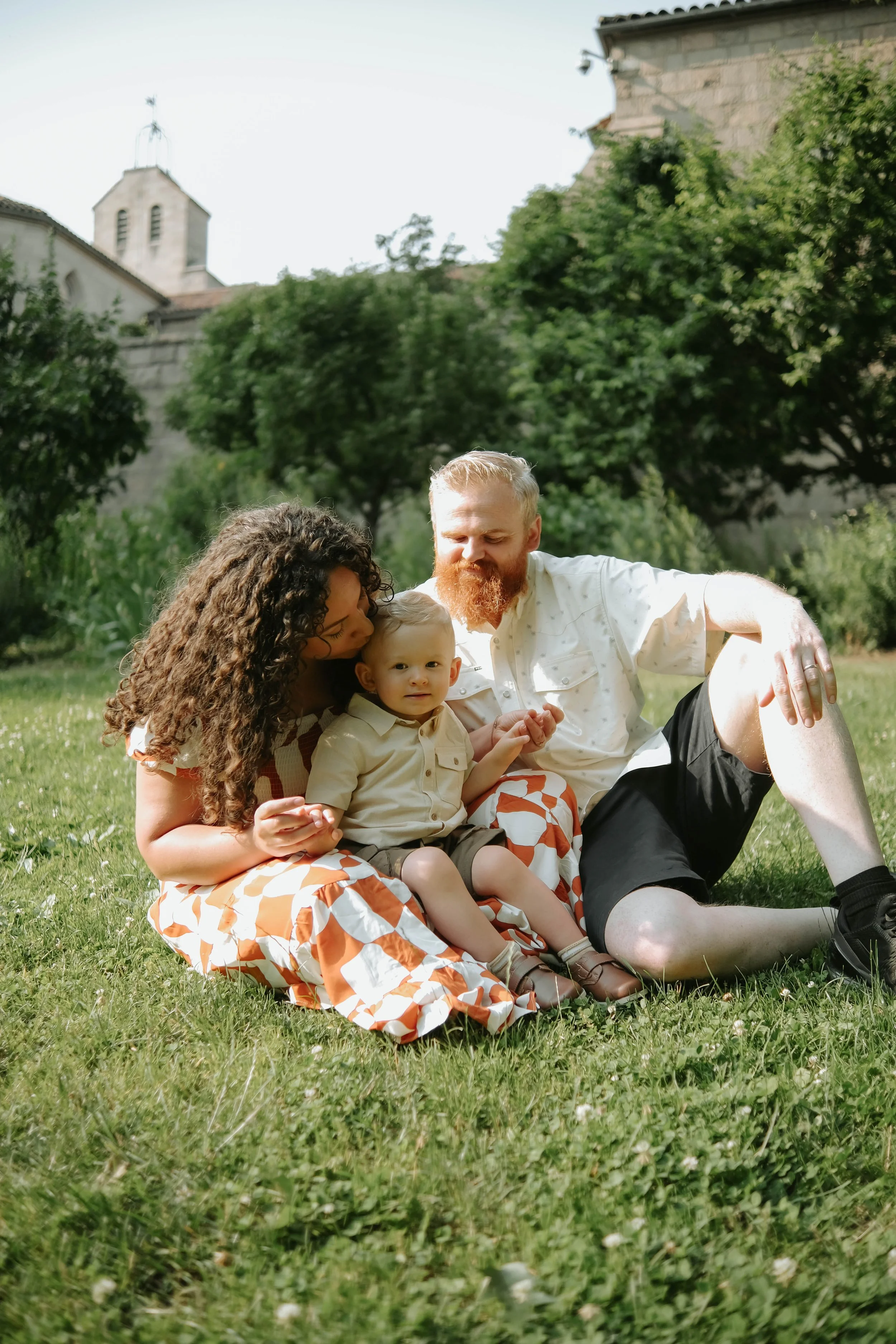 Fort Tryon Park, NYC  Family sitting on grass outdoors, with a woman, a child, and a man enjoying a sunny day surrounded by greenery and trees.