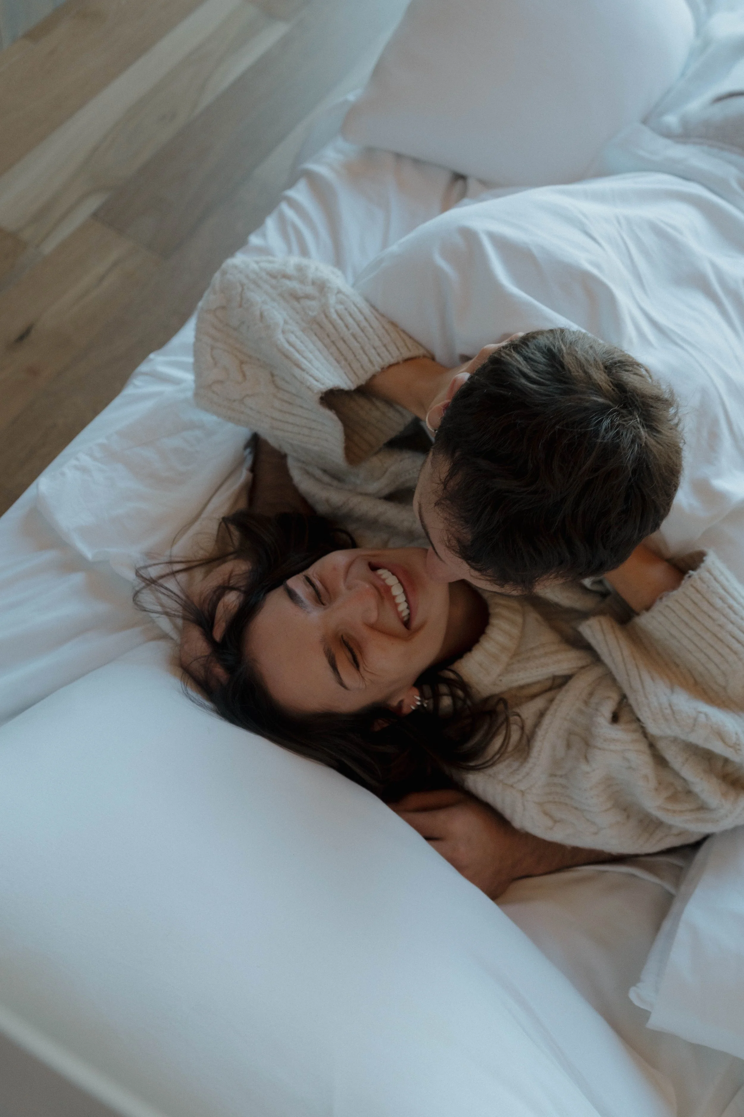 A woman and a man are cuddling and smiling on a bed with white sheets. The woman has dark hair and is wearing a beige knitted sweater, while the man has short dark hair.