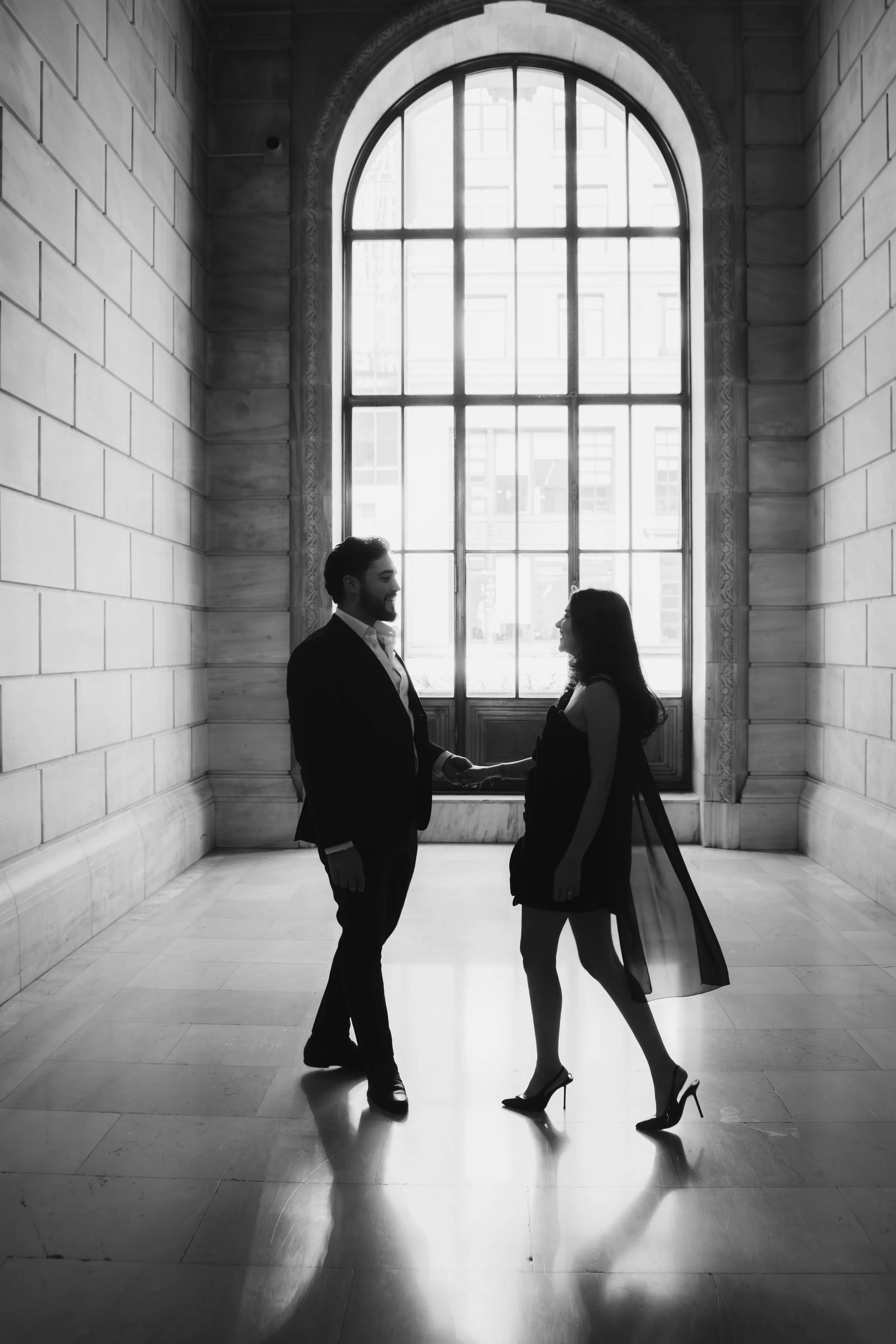 A black and white photo of a couple holding hands and facing each other inside a building with large arched window. Engagement Photoshoot at New York City Public Library with Captured by Yosi. Elegant, Emotion-filled, romantic couple.