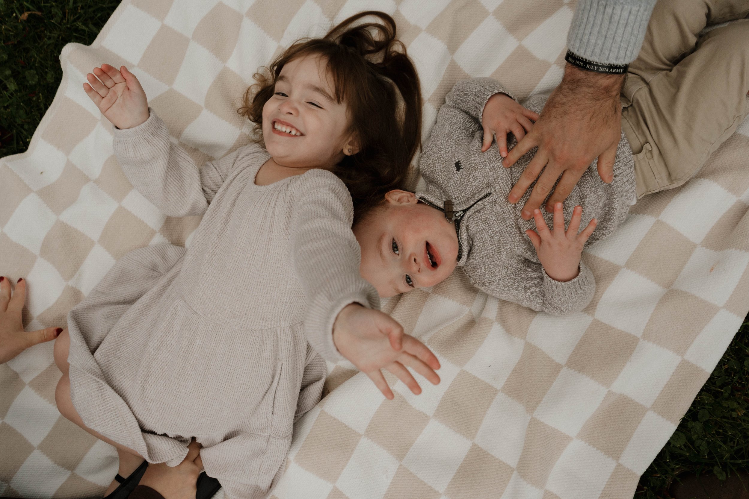 Baldwinsville, NY Photographer A smiling girl and a boy lying on a checkered blanket outdoors, with a hand touching the boy and others reaching towards the girl.