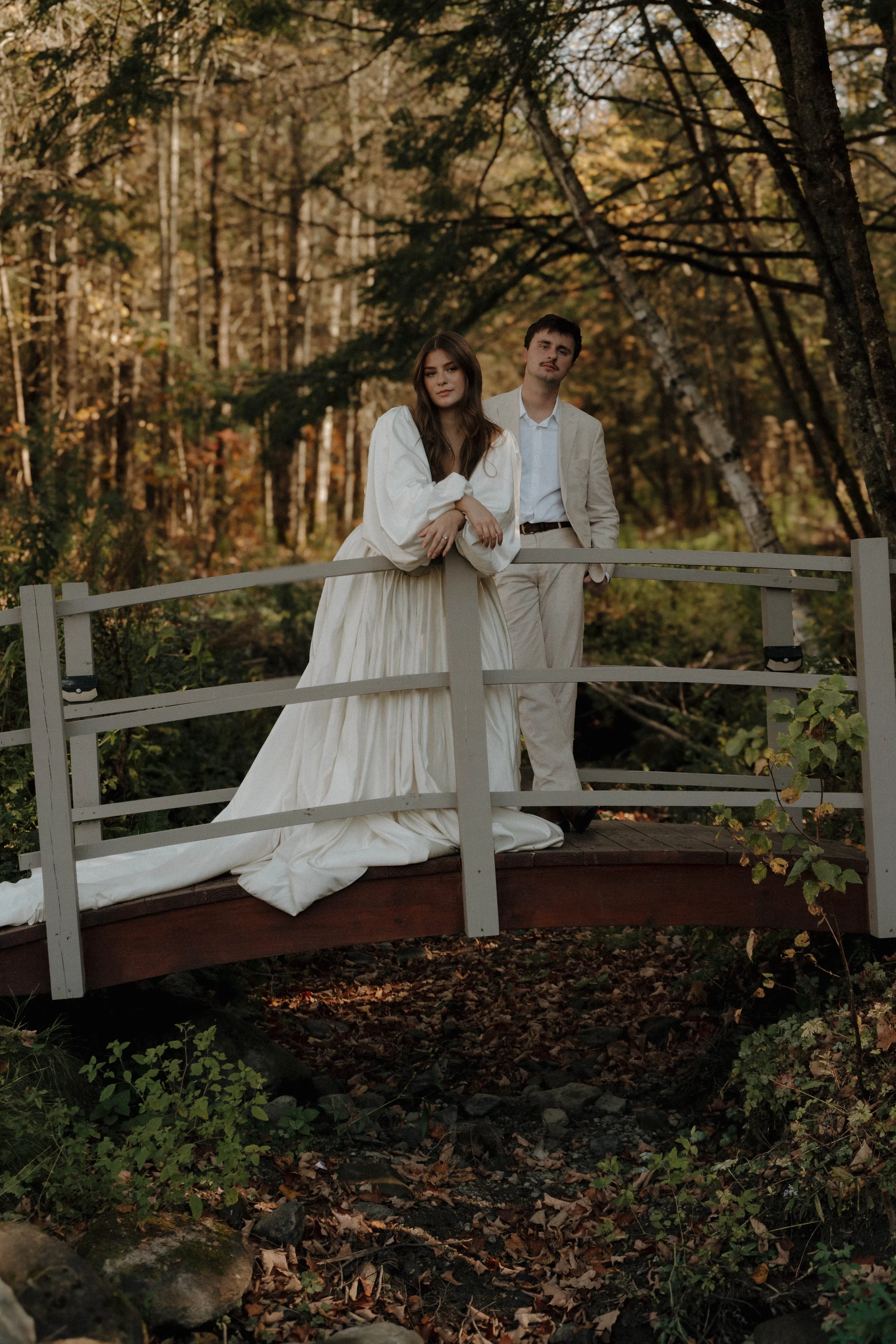 A woman in a white vintage wedding dress and a man in a light suit standing on a small wooden bridge in a forest with autumn foliage.