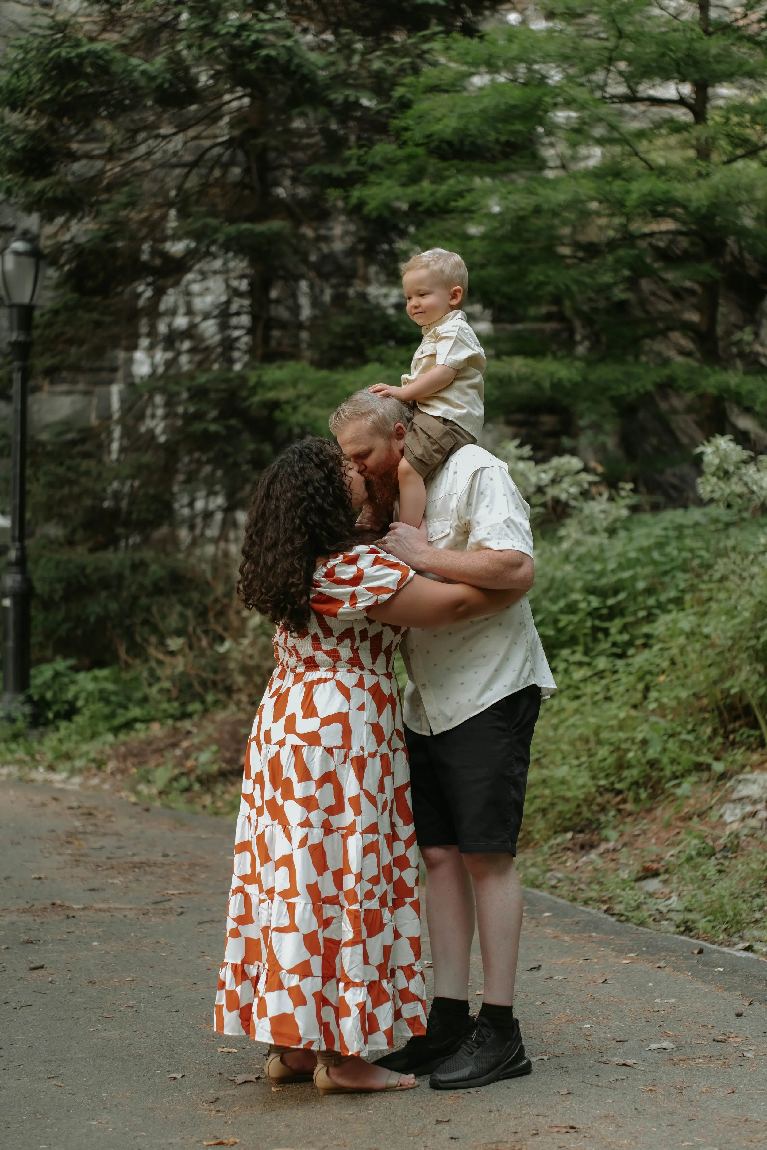 Fort Tryon Park, NYC A family of three sharing a kiss outdoors, with a woman and man embracing and a boy sitting on the man's shoulders, surrounded by greenery.