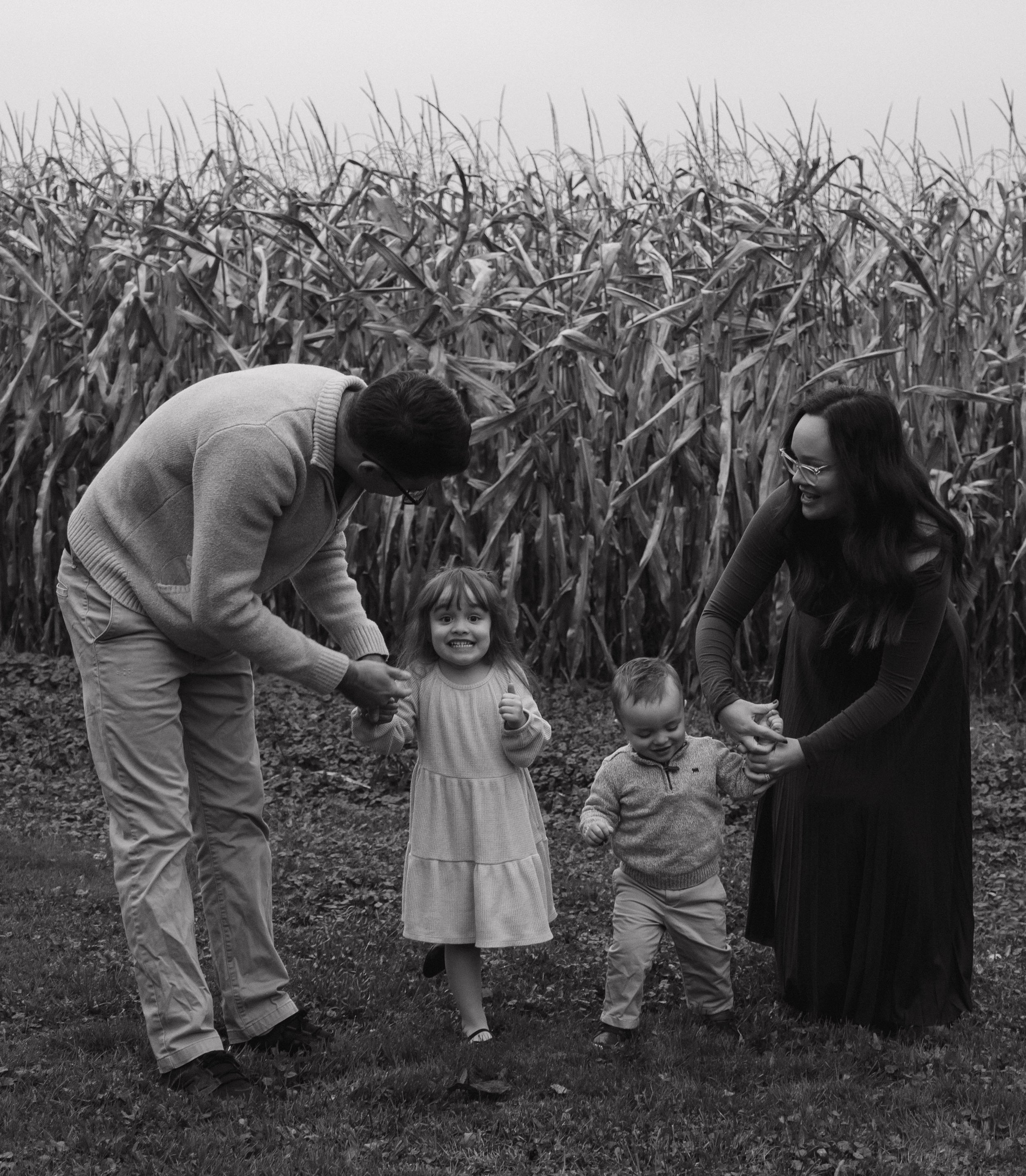 Baldwinsville, NY Photographer A family of four, including a man, woman, young girl, and young boy, holding hands and playing in a field near a cornfield.