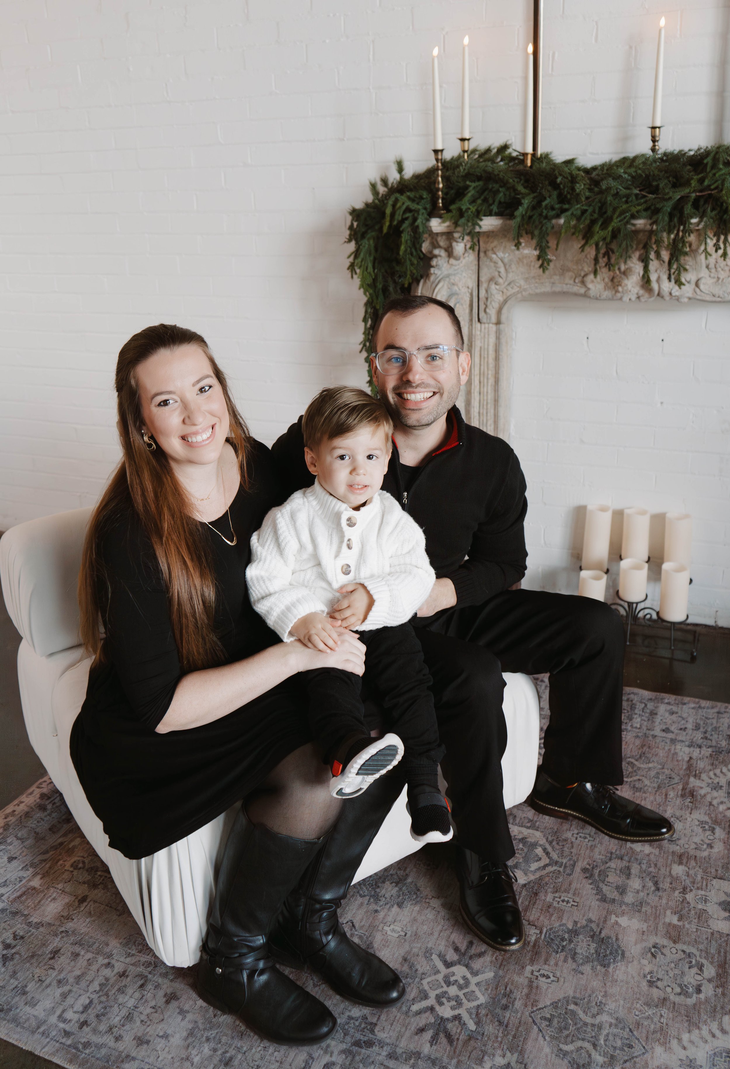 Pose Studio Photographer, A family of three sitting on a white sofa in front of a decorated fireplace with candles and greenery, smiling at the camera.