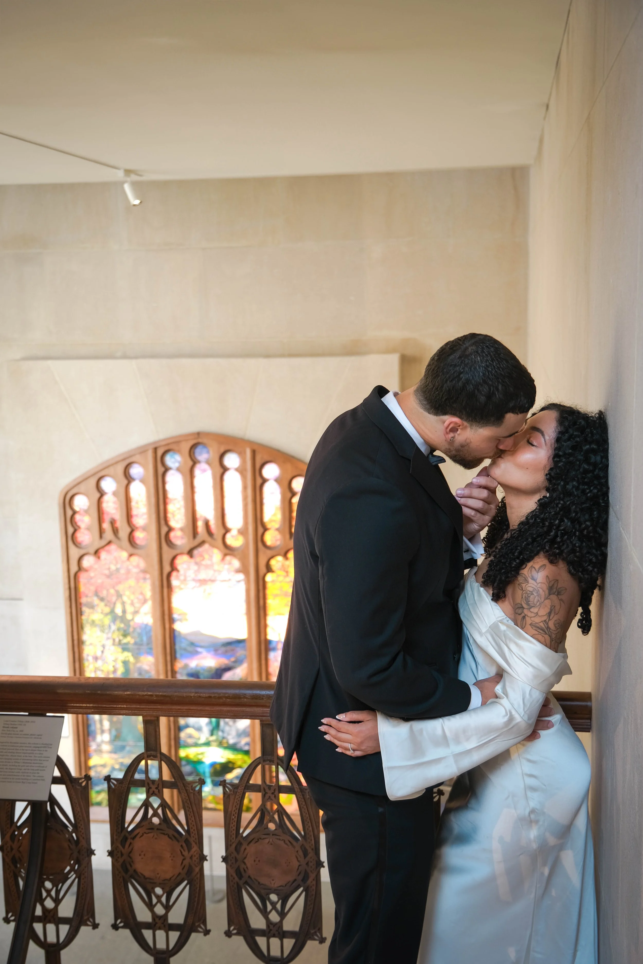 The Met Engagement Shoot, Couple in wedding attire sharing an intimate moment, with the man gently holding the woman's face while they are about to kiss, against a luminous background of stained glass windows.
