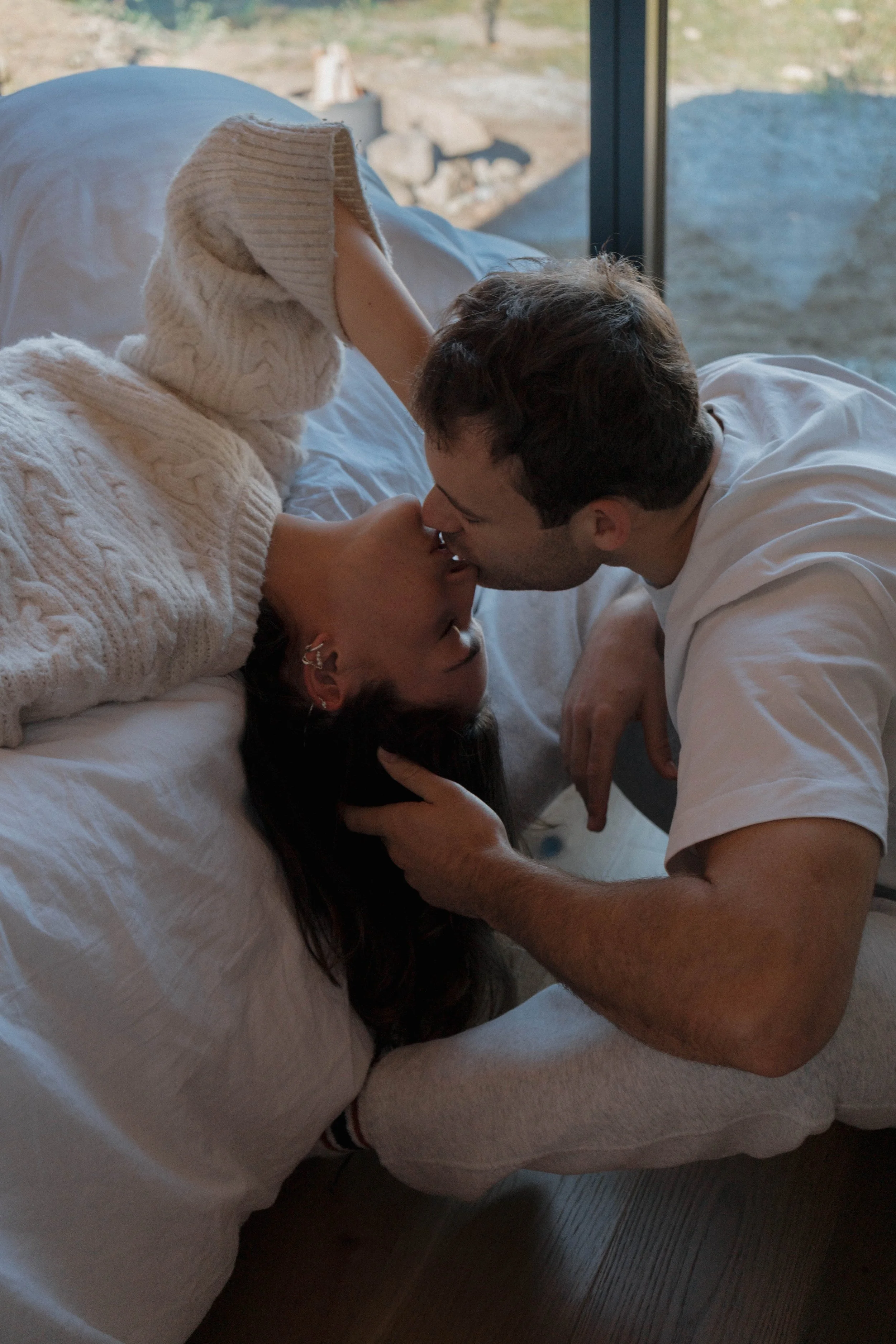 A couple sharing a kiss on a bed indoors with a window in the background.