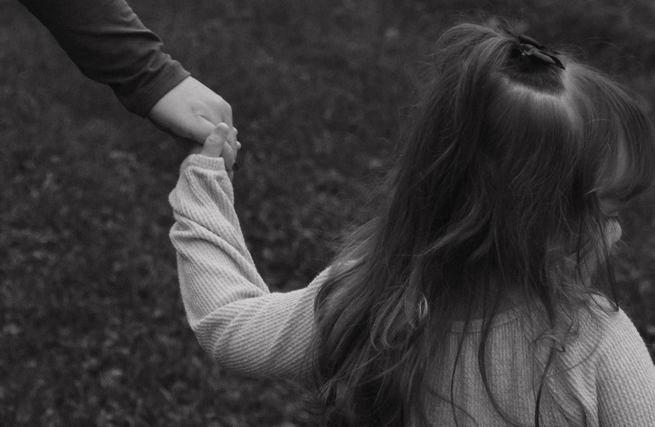 Baldwinsville, NY Photographer A young girl with long hair tied with a bow, holding hands with an adult, outdoors on a grassy area.