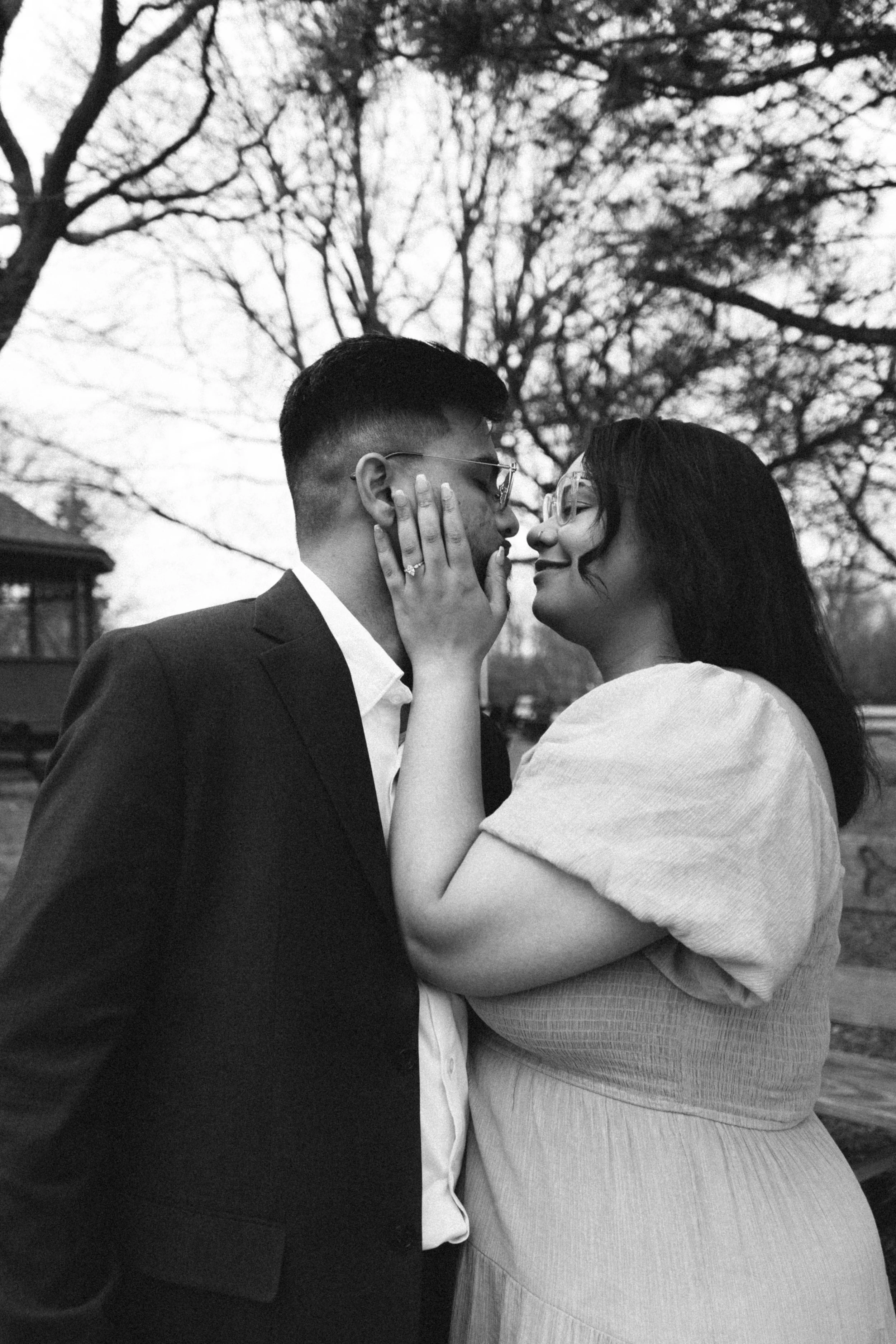 A black and white photo of a couple with light skin, standing close and smiling at each other outdoors. The woman her hands on the man's face, and they are about to kiss, with leafless trees and a house in the background.