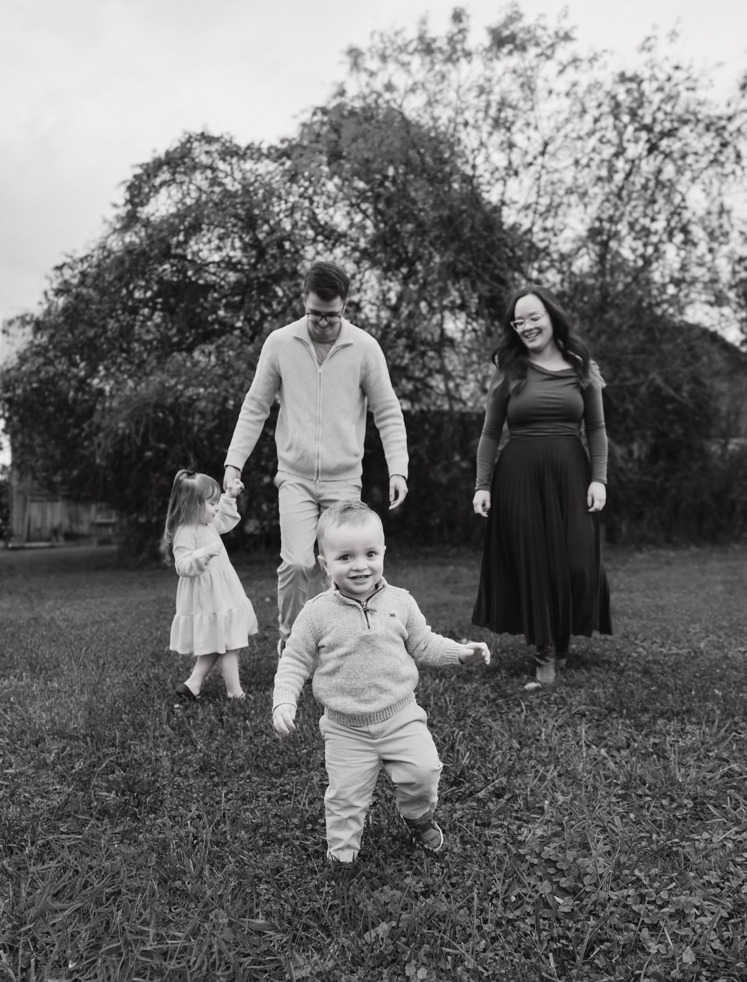 Baldwinsville, NY PhotographerA happy family of four, including two young children, playing outdoors on a grassy field with trees in the background. The children are smiling and one is walking towards the camera while the other holds an adult's hand.