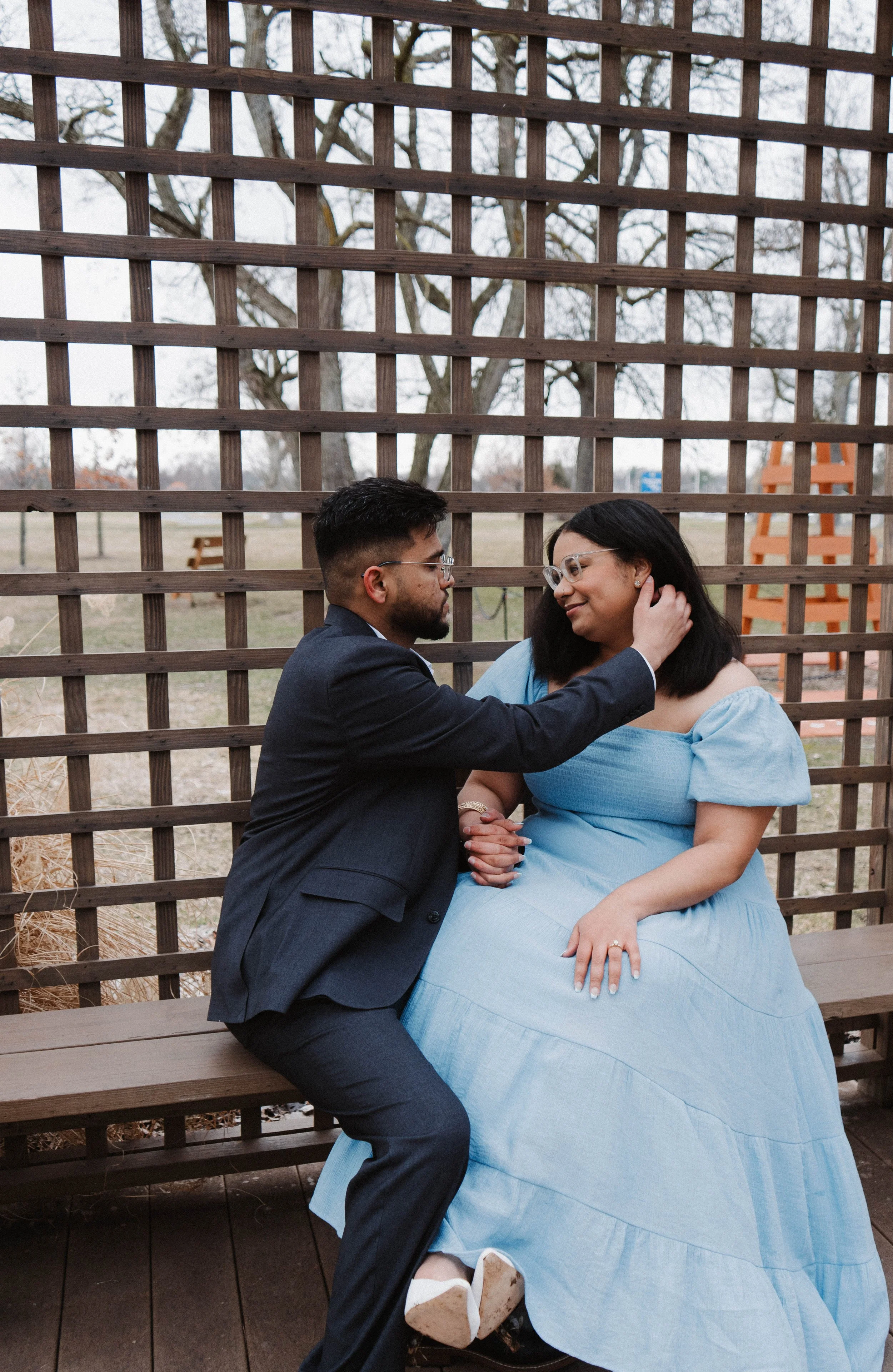 A couple sitting on a wooden bench outside, with the man gently touching the woman's face. They are looking at each other affectionately, with a wooden lattice behind them and leafless trees in the background.