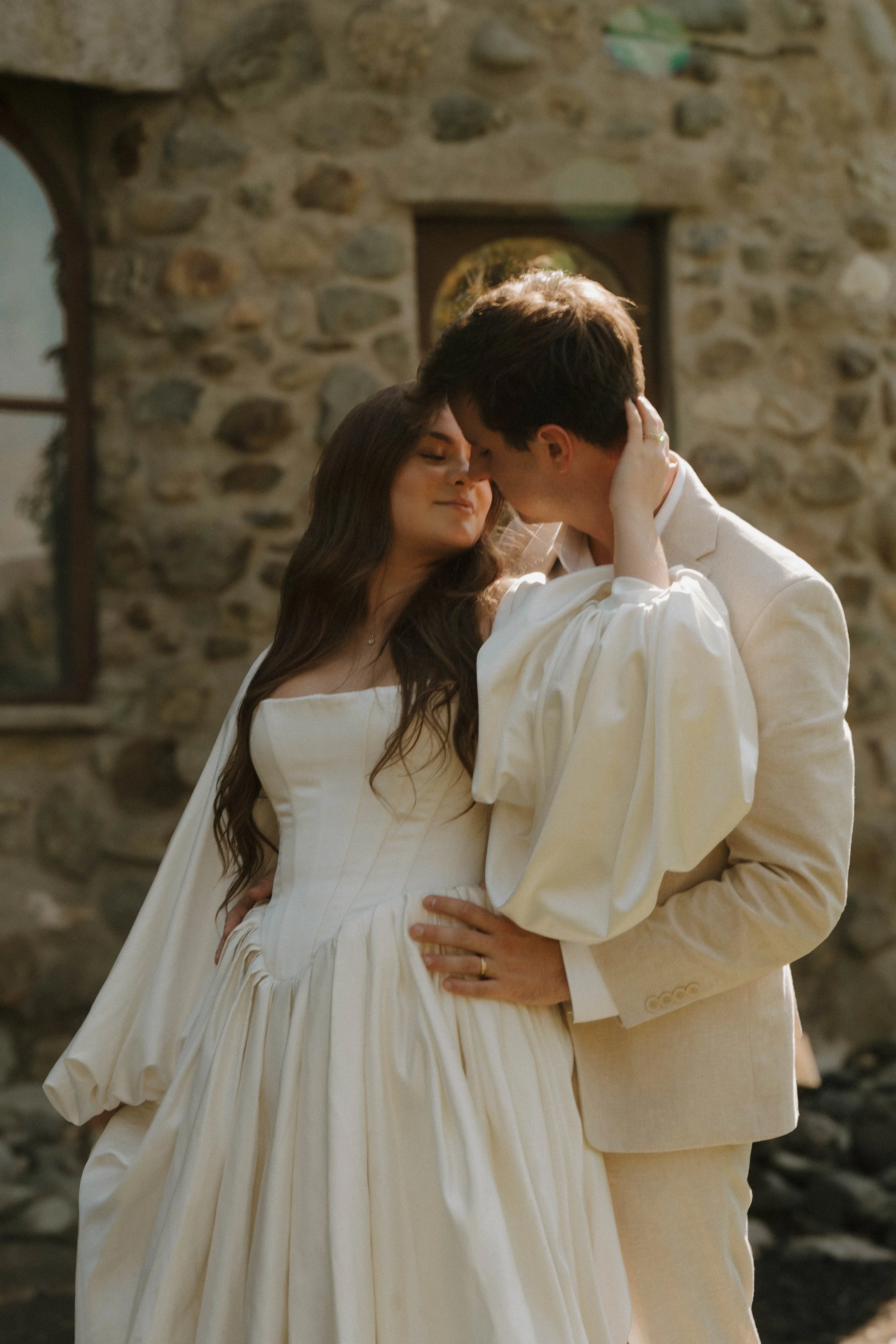 A couple is dancing closely in wedding attire, with the woman in a white gown and the man in a cream suit, in front of a rustic stone wall.