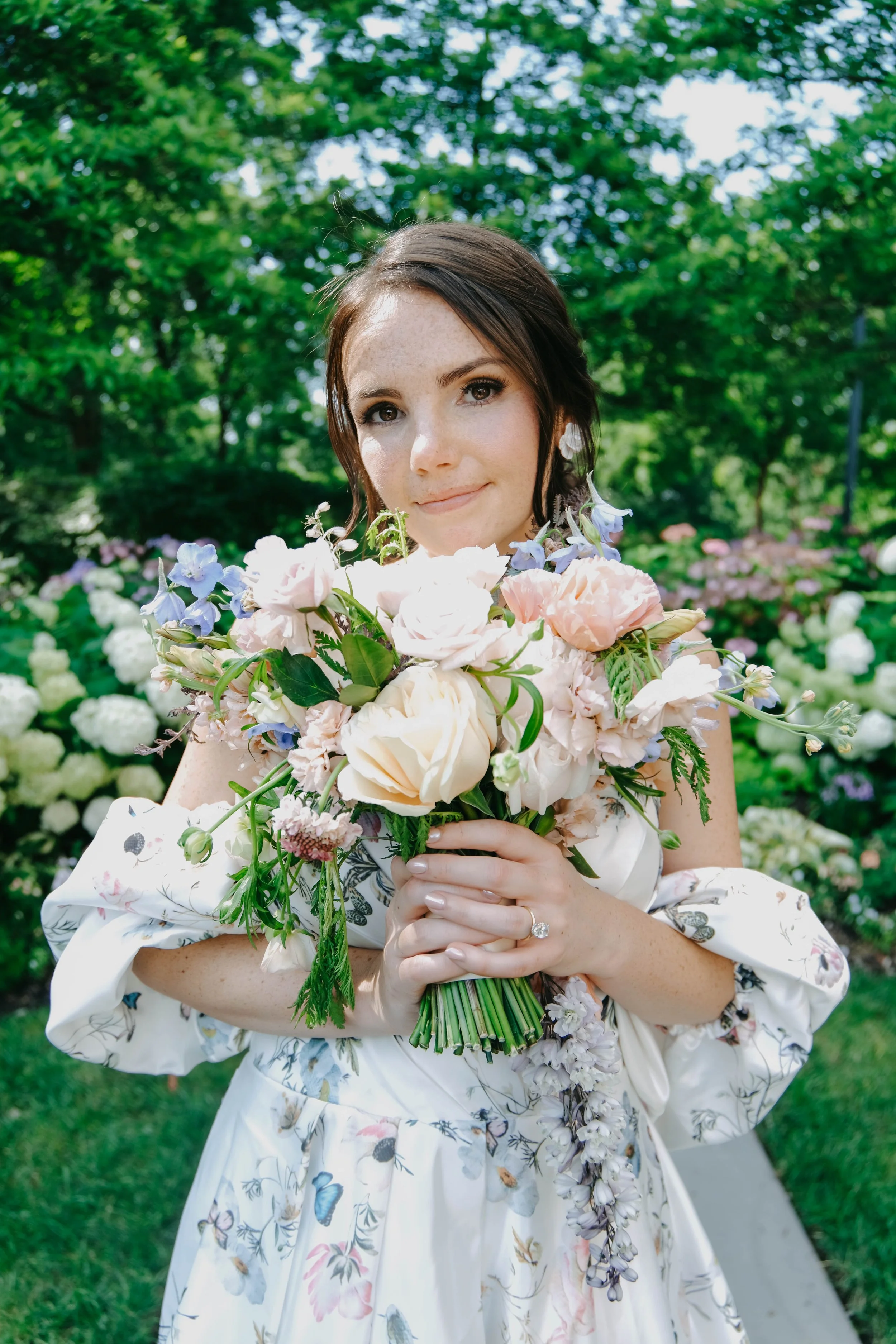A woman in a floral dress holding a bouquet of flowers outdoors with greenery in the background.
