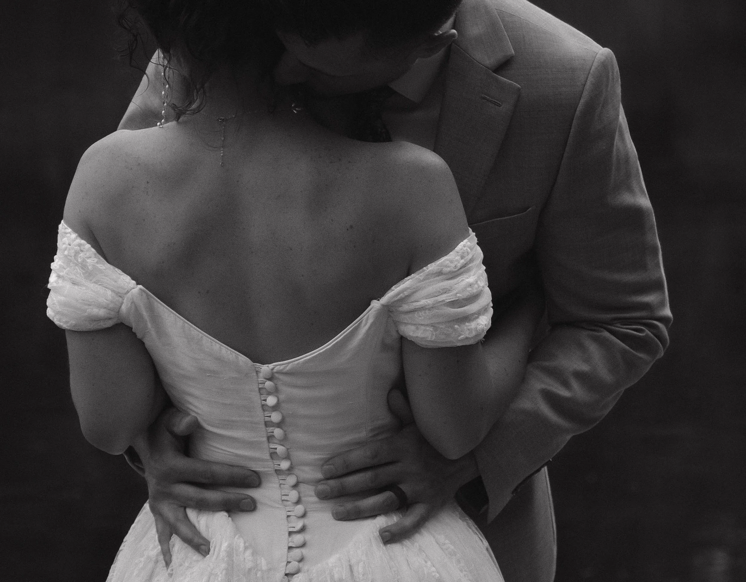 A black and white photo of a couple embracing, with the woman wearing an off-shoulder dress with buttons down the back, and the man in a suit jacket, holding her around the waist.
