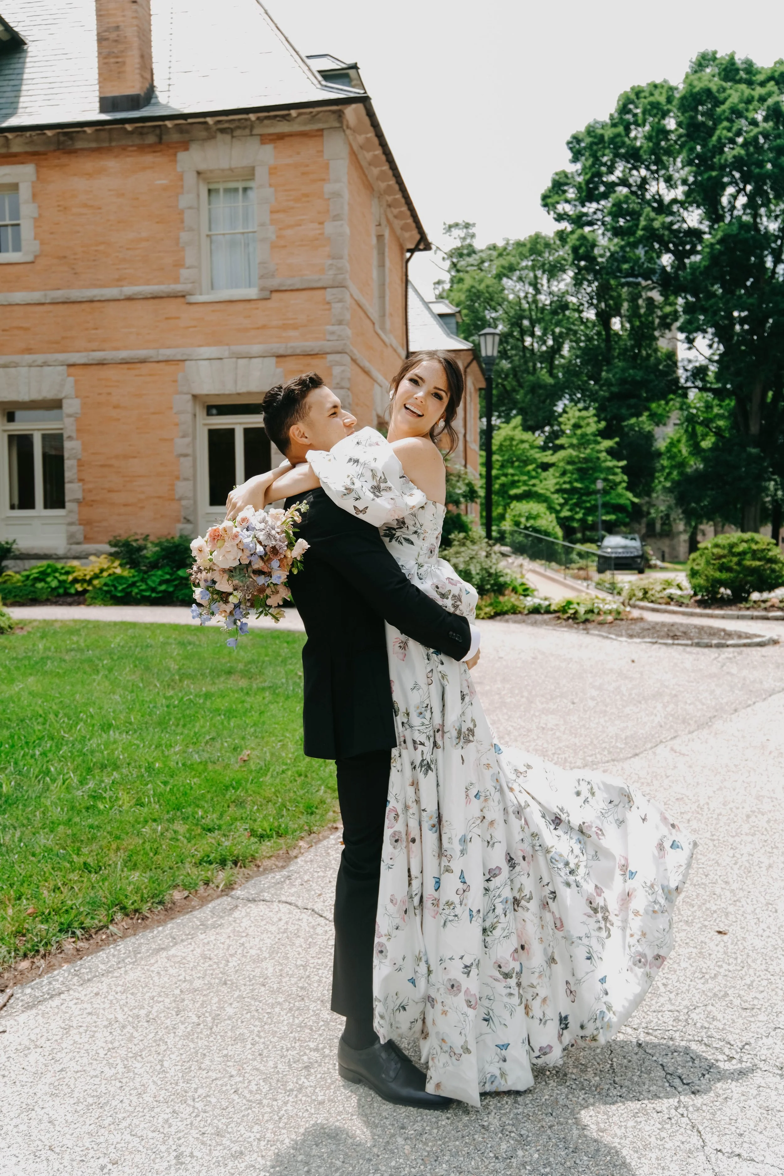 A happy bride and groom, with the groom in a black suit and the bride in a white floral wedding dress, standing outdoors in front of a brick building, celebrating their wedding day.