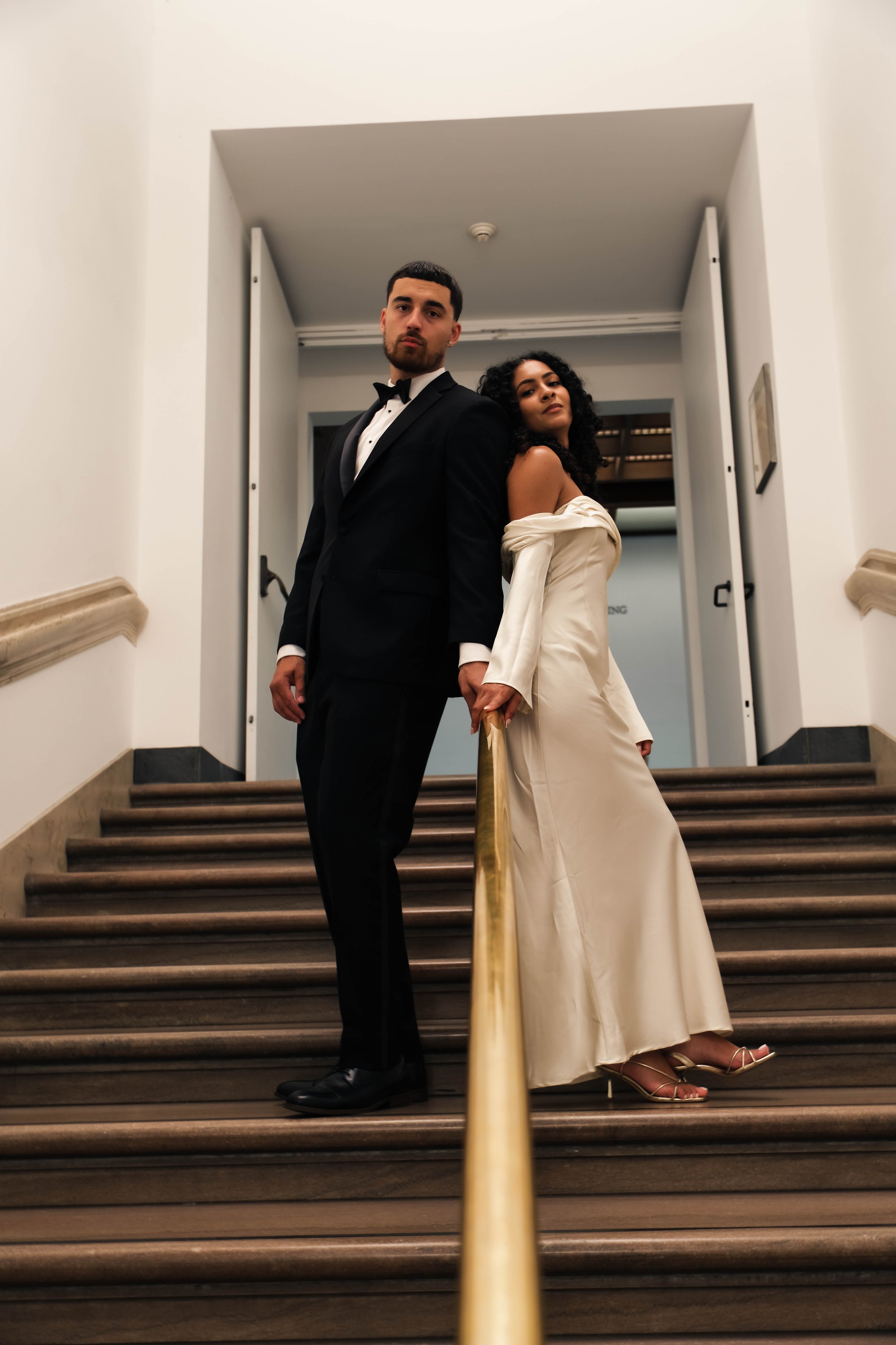 The Met Engagement Shoot  A couple in formal attire standing on a staircase, holding hands behind a gold-colored handrail, in an elegant indoor setting.