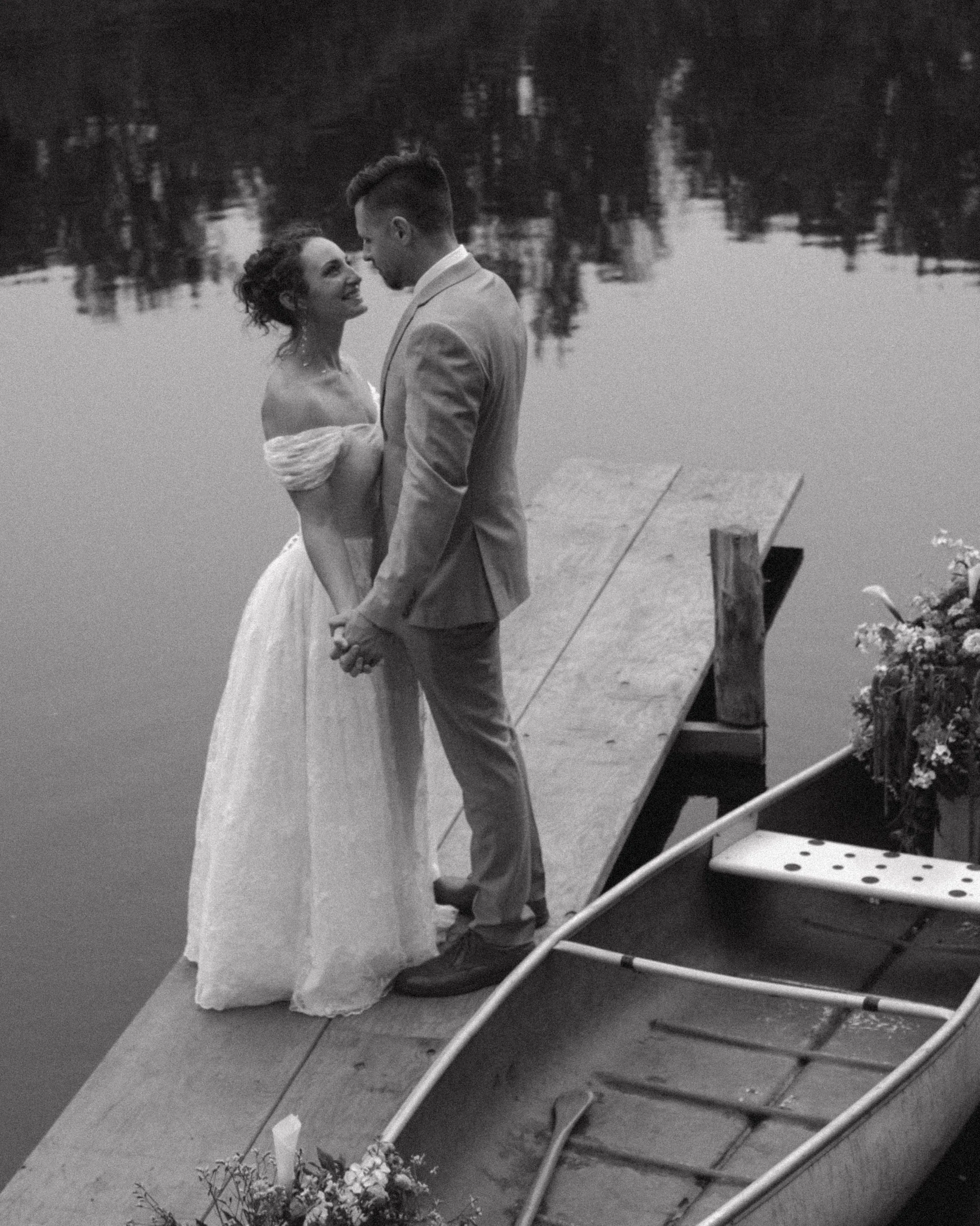A black and white photo of a bride and groom standing on a wooden dock by a lake, holding hands, gazing at each other, with flowers beside the boat dock.