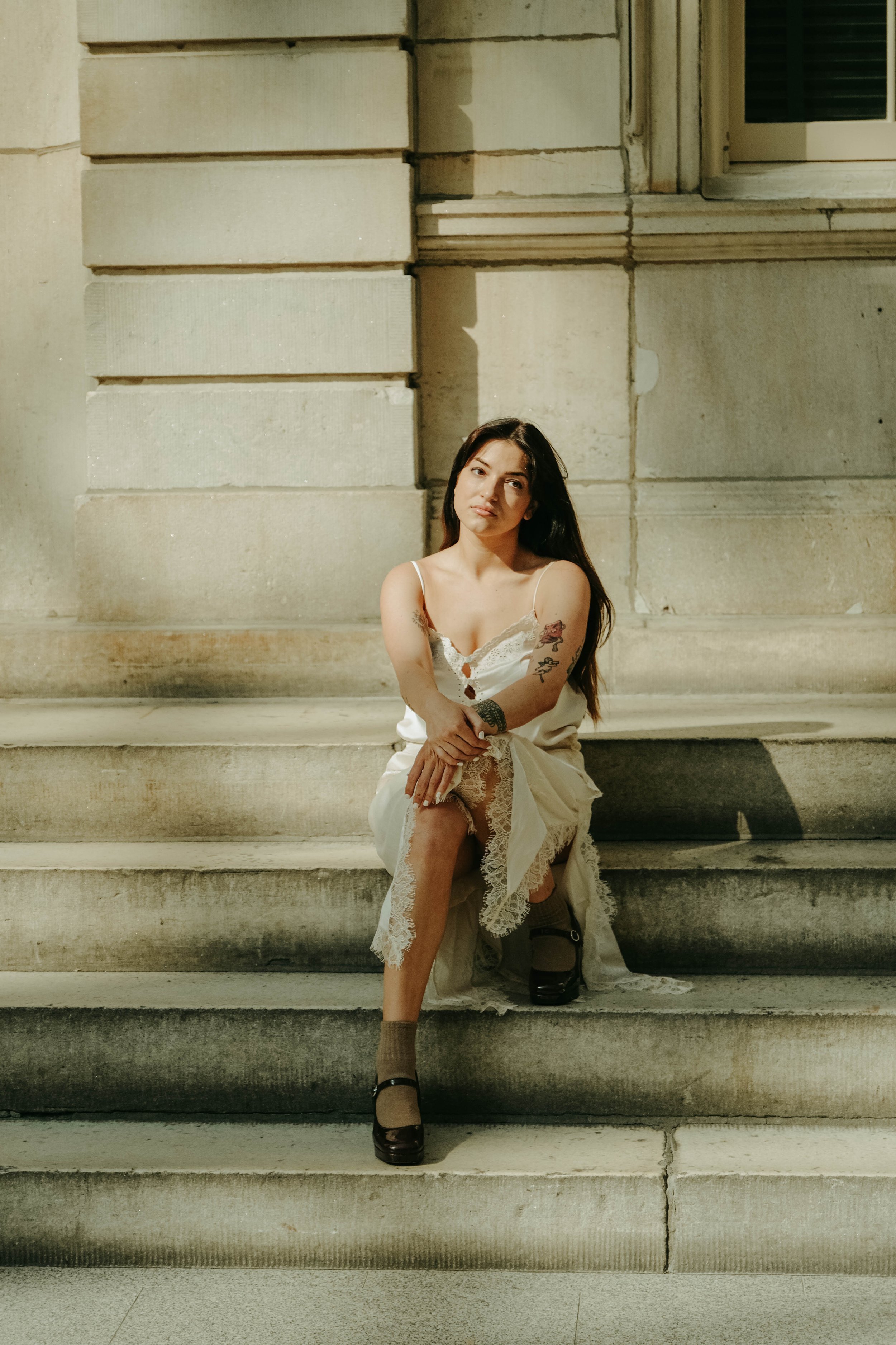 The Met Museum, A young woman with tattoos, long dark hair, and wearing a white slip dress with lace trim, sits on stone steps in front of a stone building, illuminated by sunlight.