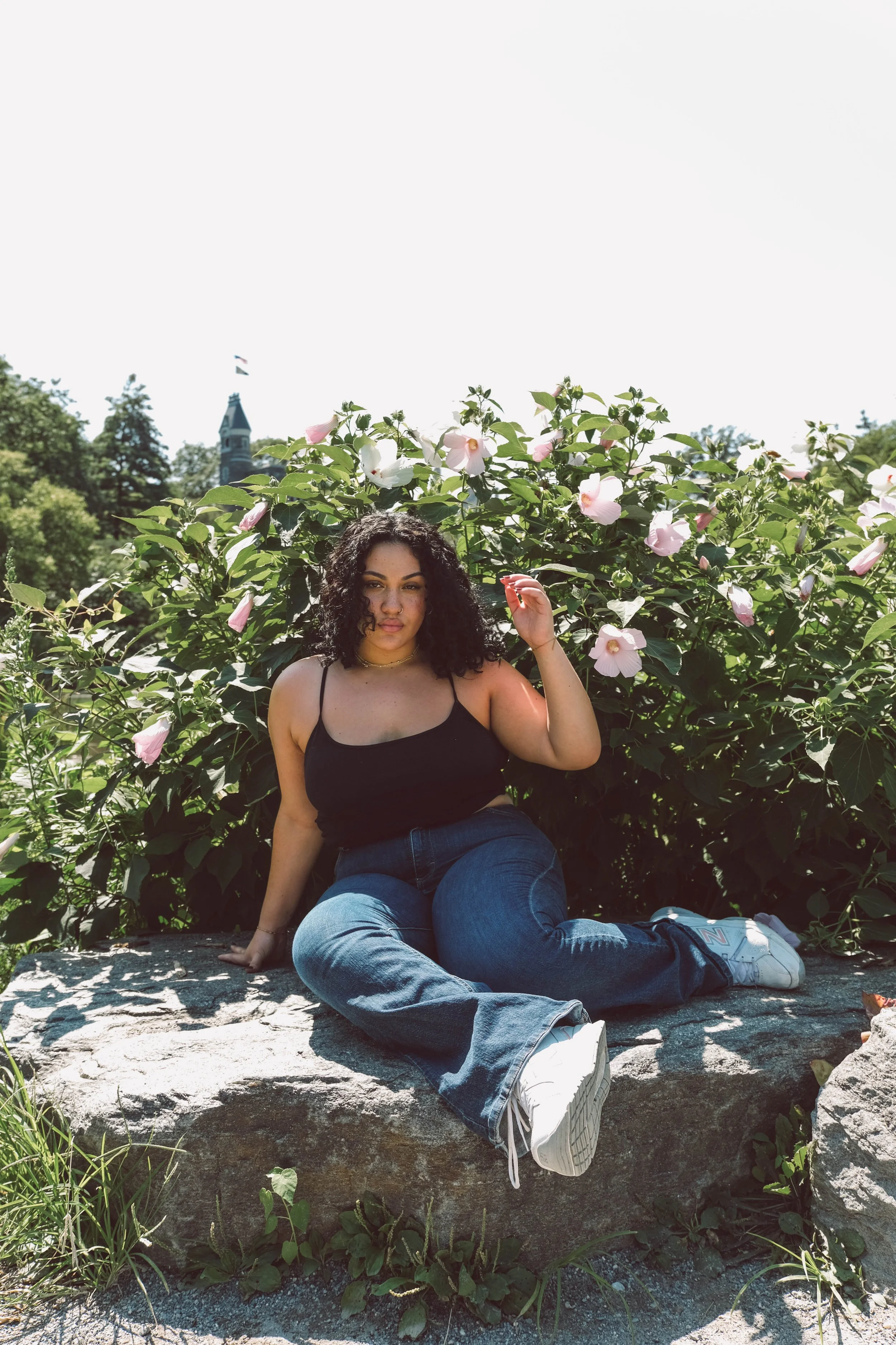 Central Park, a woman with curly dark hair, wearing a black spaghetti strap top and blue jeans, sitting on a large rock in front of blooming pink and white flowers, with greenery and a building with a pointed roof in the background, on a sunny day.