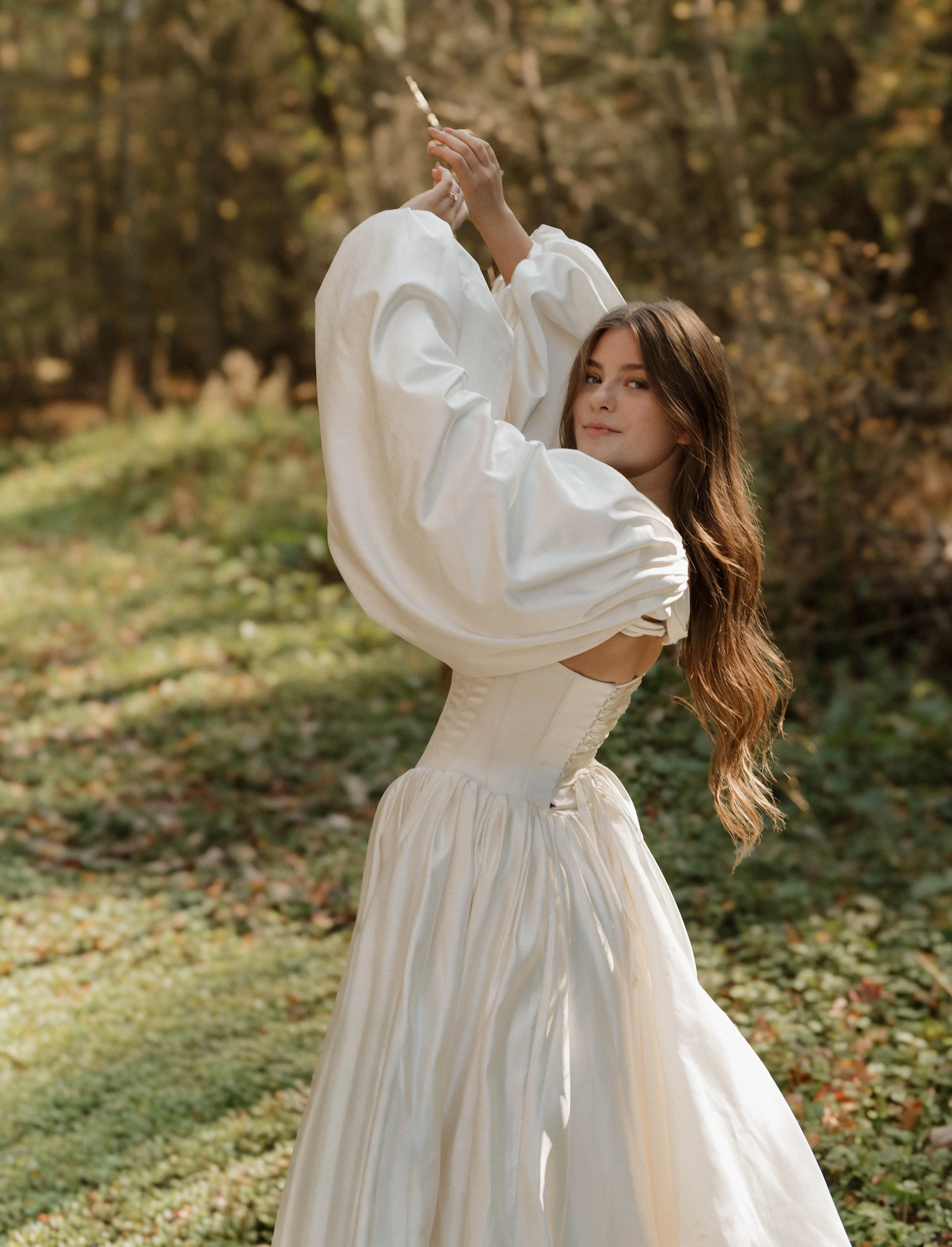 Vermont, Wedding, A woman with long, wavy hair wearing a vintage-style white dress with puffed sleeves and a corset is outdoors in a wooded area during autumn.
