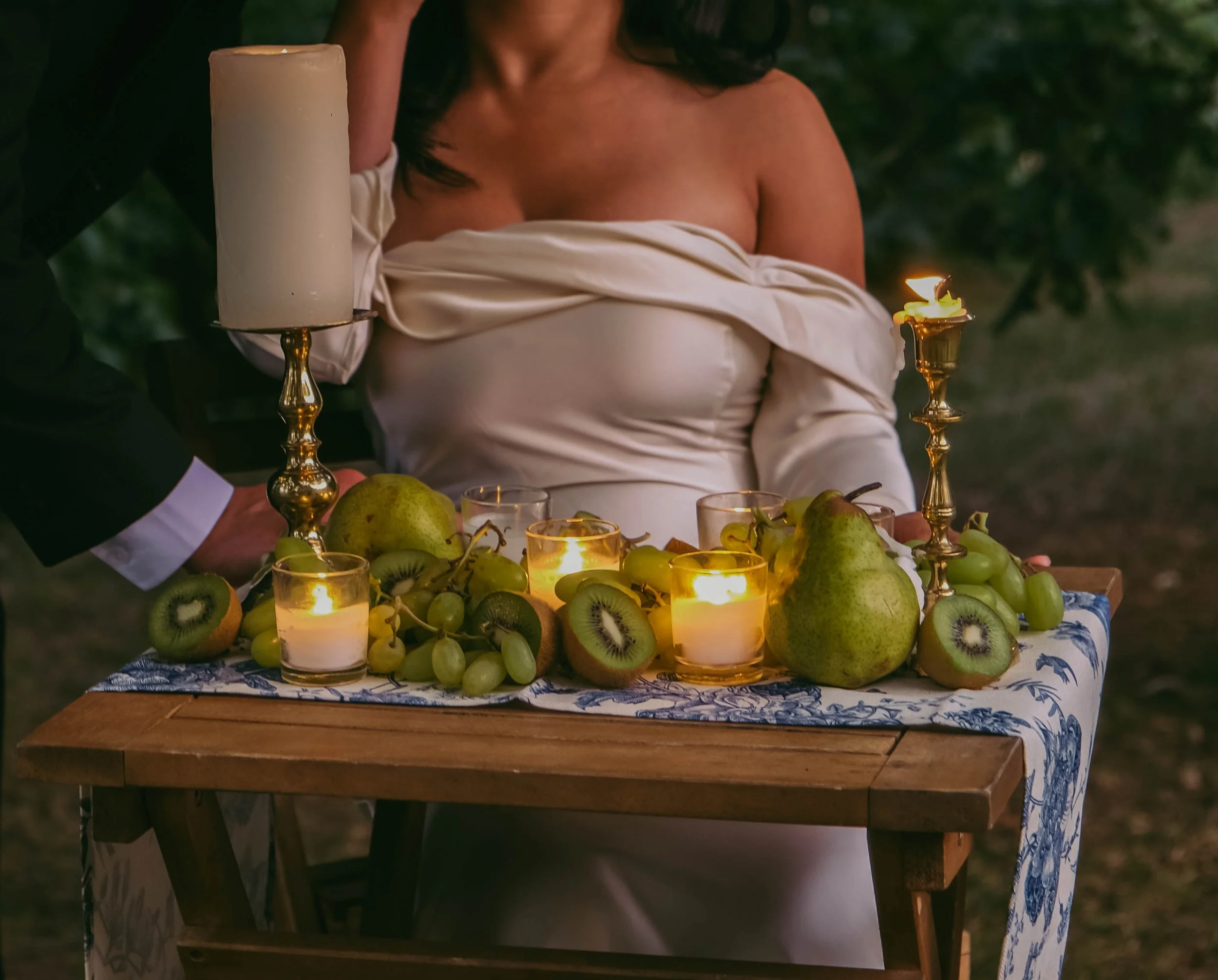 Potsdam, NY Photographer A woman in a white dress standing behind a table decorated with candles, fruits including pears, kiwis, and grapes, and a blue and white cloth, during an outdoor evening setting.
