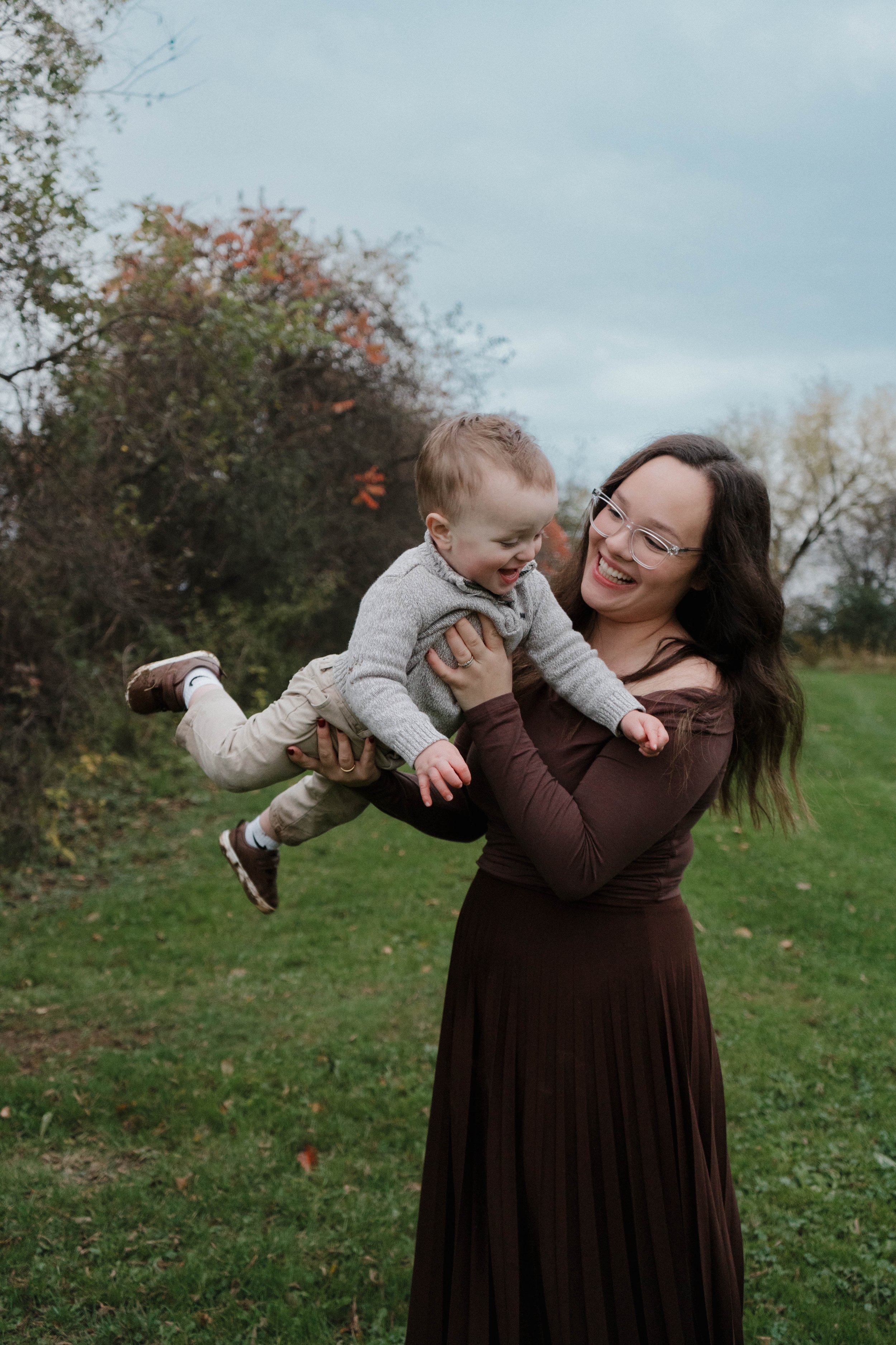 Baldwinsville, NY Photographer A woman with glasses lifting a young boy in an outdoor park with trees and grass, both smiling and enjoying the moment.