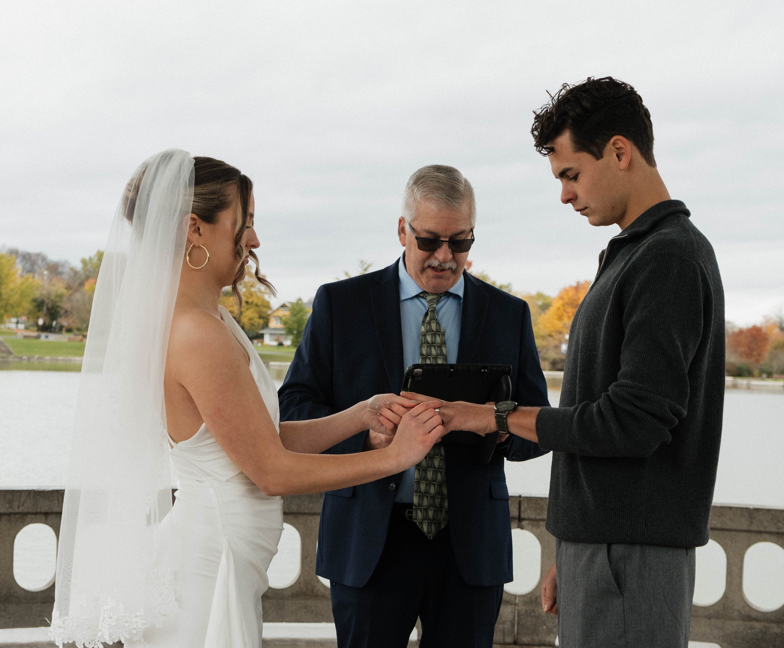 A bride and groom exchange rings outdoors by a lake with trees showing fall colors, as an officiant presides.
