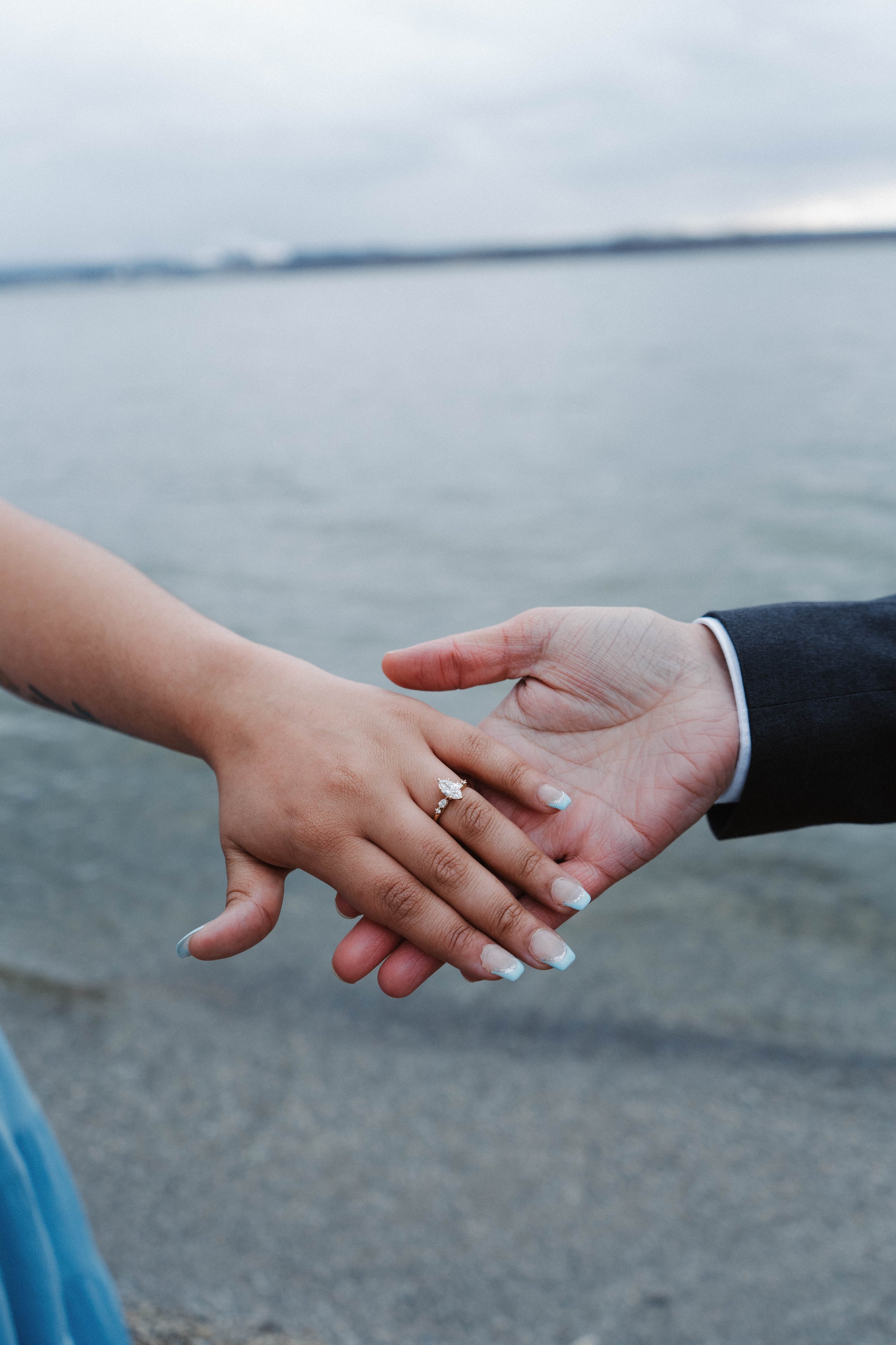 A person wearing a wedding ring holding hands with another person dressed in a suit near a body of water.
