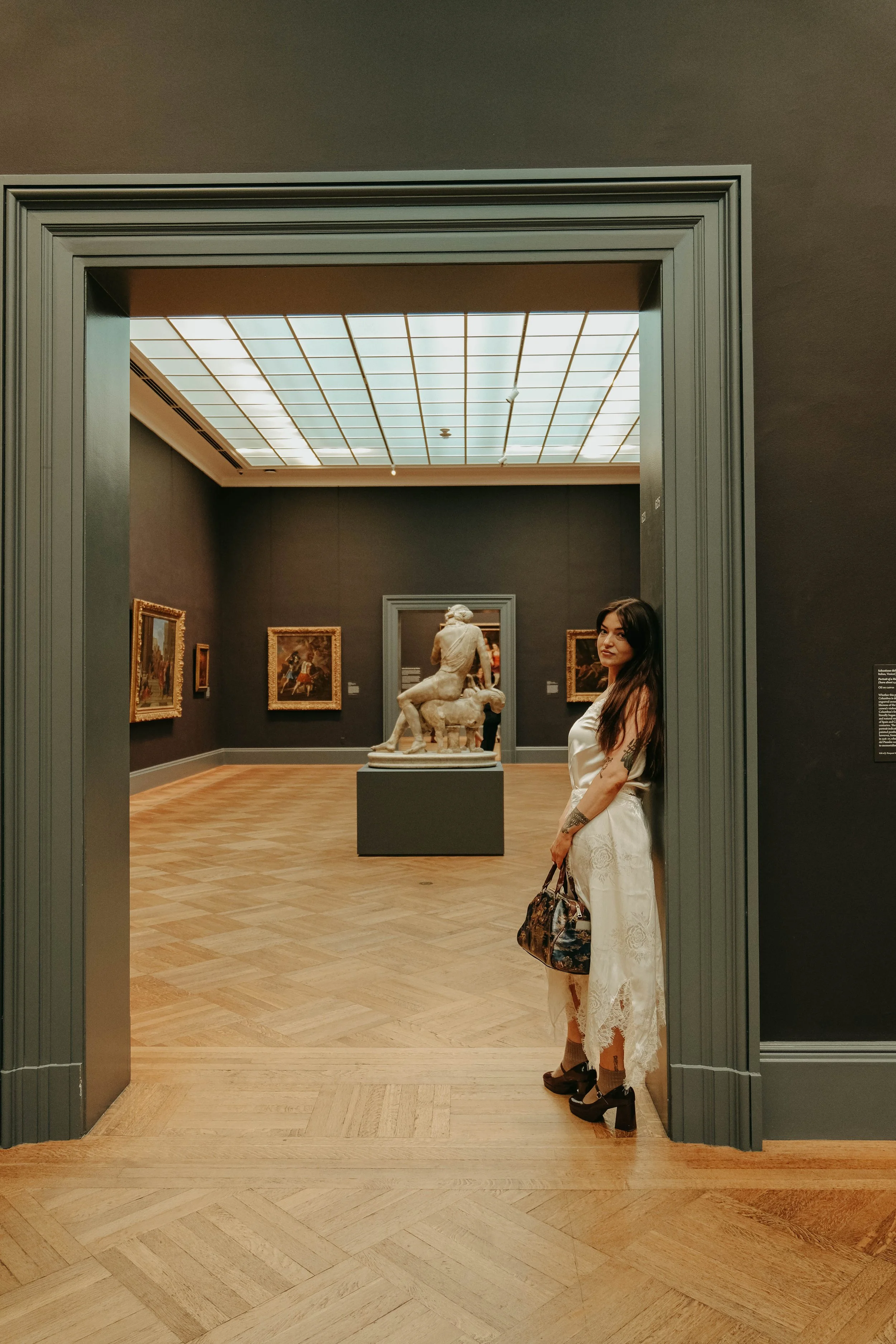 The Met Museum, Woman in white dress standing inside a museum, looking at the camera, with sculptures and paintings displayed on dark walls behind her.