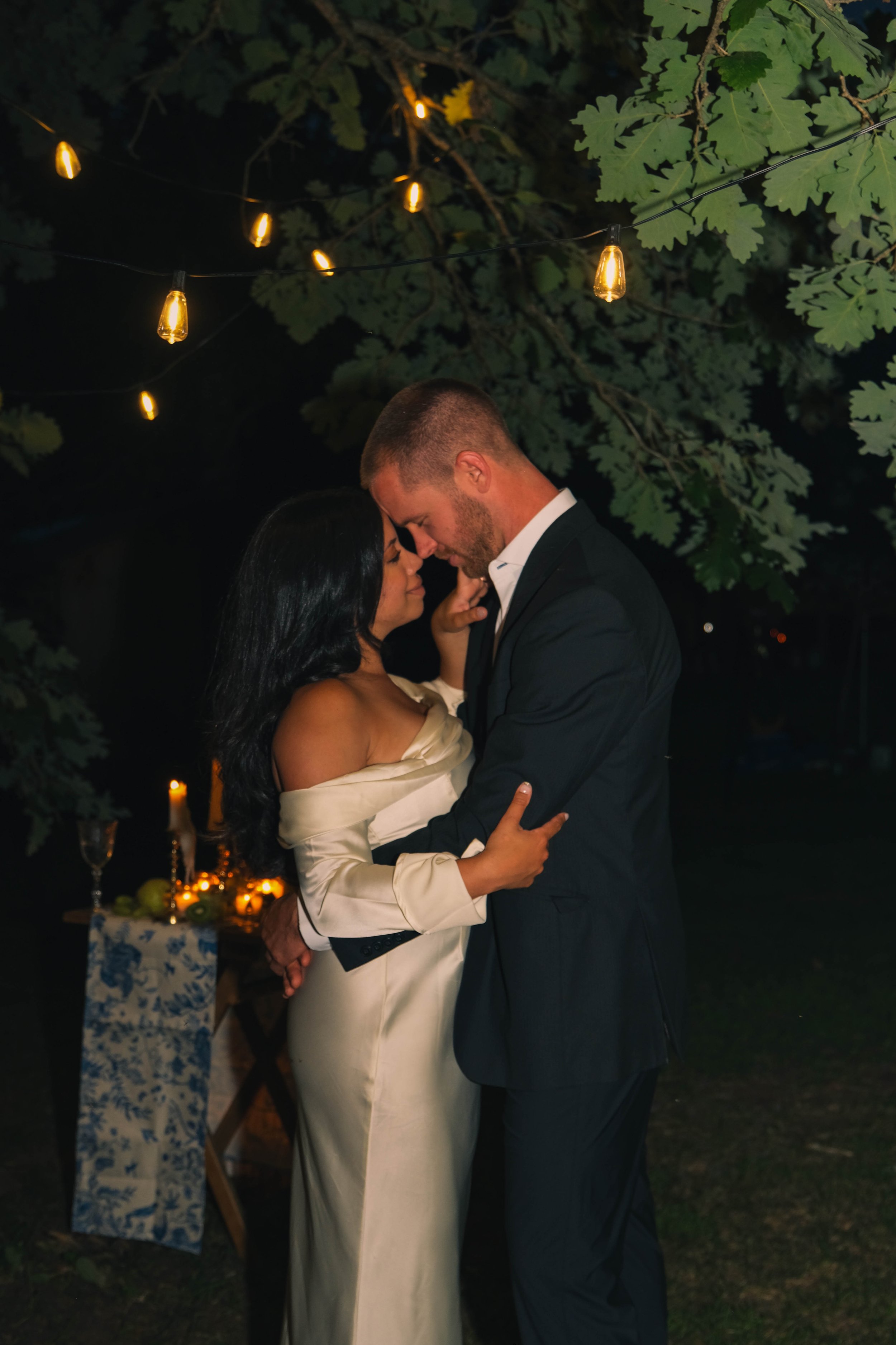 A romantic night scene of a couple dancing outdoors under string lights, with a table and candles in the background.