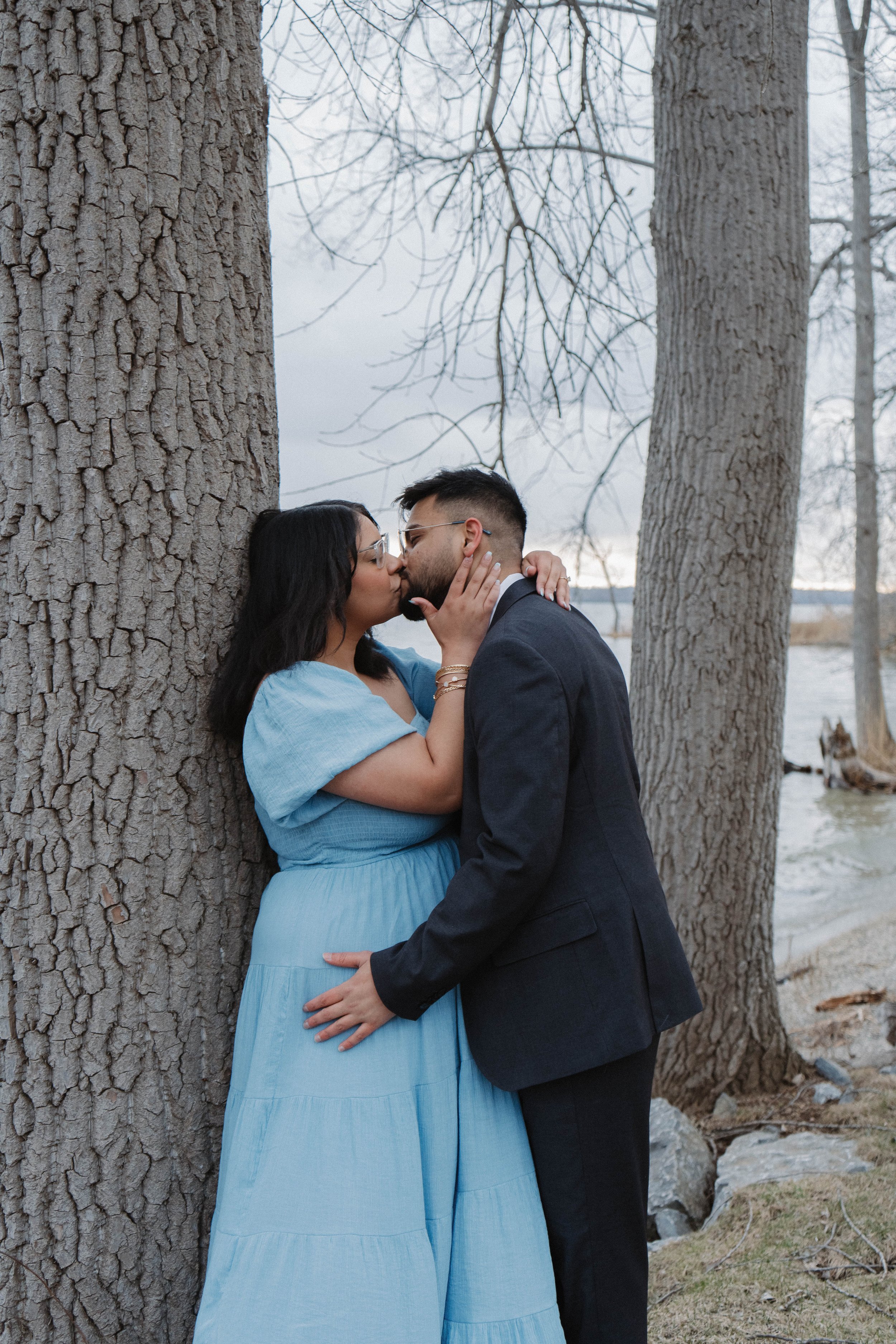 A couple sharing a kiss outdoors near a large tree, with a lake and leafless trees in the background.