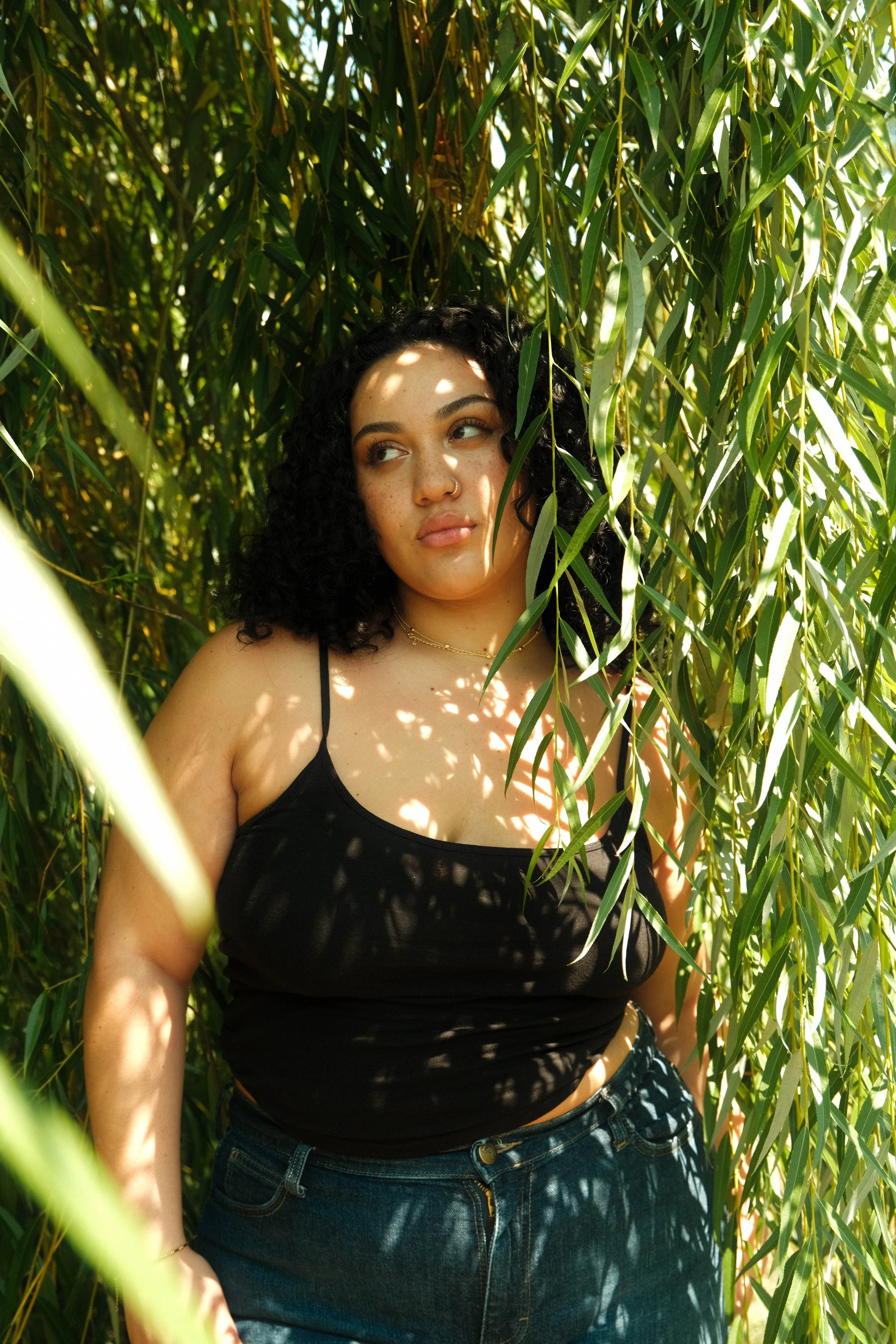 Central Park, Young woman with curly black hair, wearing a black tank top and jeans, standing among green plants and leaves with sunlight filtering through.