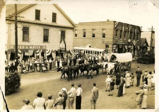Historical black-and-white photo of a town street parade featuring a horse-drawn wagon, people lining the sidewalk, and vintage storefronts. Wilton NH